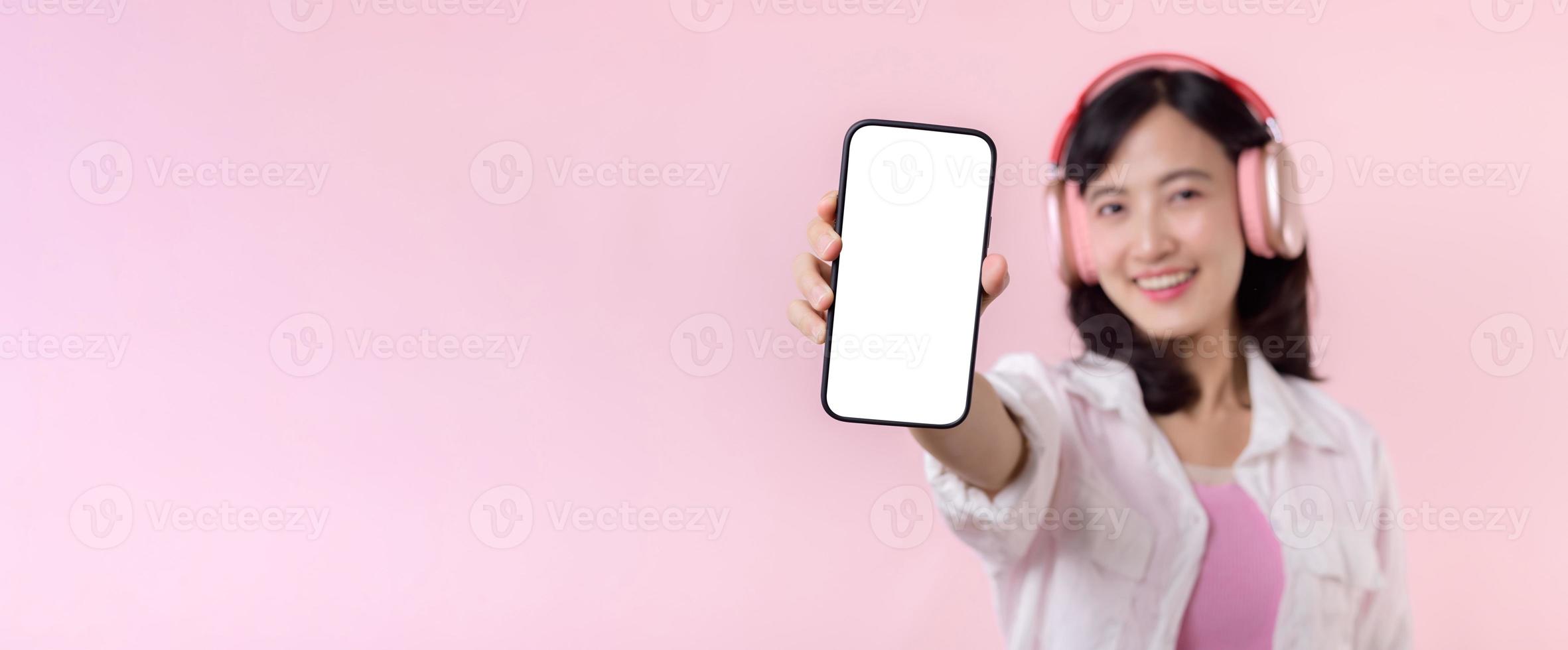 feliz alegre sorridente ásia mulher com sem fio fones de ouvido mostrando em branco tela Móvel telefone ou Novo Smartphone música inscrição propaganda brincar isolado em Rosa estúdio fundo. foto