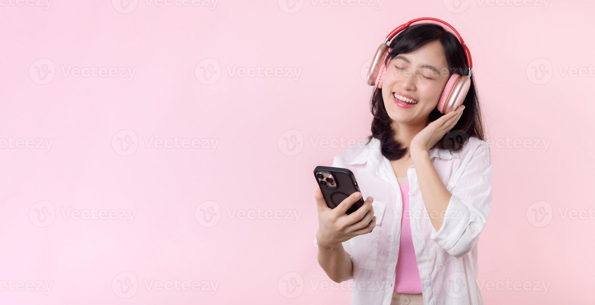 retrato alegre jovem ásia mulher apreciar ouvindo audio de Smartphone música inscrição contra Rosa. feliz sorridente fêmea pessoa com fone de ouvido. som, lazer, estilo de vida, tecnologia conceito foto