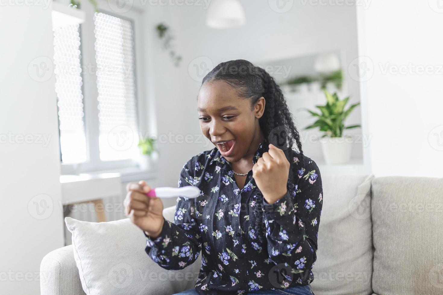 jovem mulher olhando às gravidez teste dentro felicidade. finalmente grávida. atraente Preto mulheres olhando às gravidez teste e sorridente enquanto sentado em a sofá às casa foto