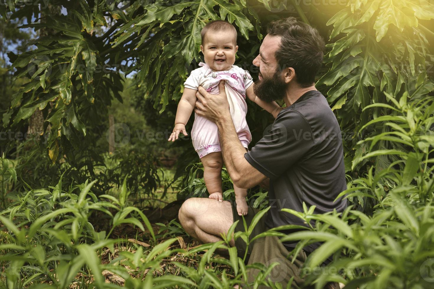 uma bebê menina parece às a Câmera sorridente, dela pai detém. ao ar livre vida foto