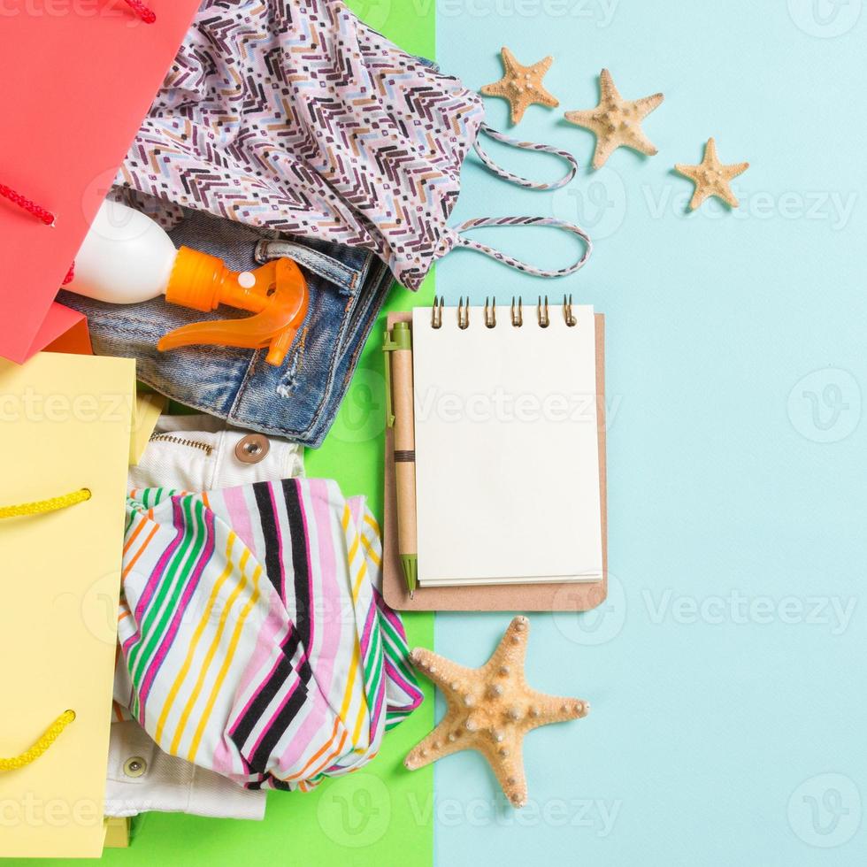 conceito de verão de sacolas coloridas cheias de roupas. sacolas de presente com camiseta, shorts jeans, notebook e frasco de protetor solar. conceito de lista de desejos de verão. espaço de cópia de vista superior no fundo azul foto