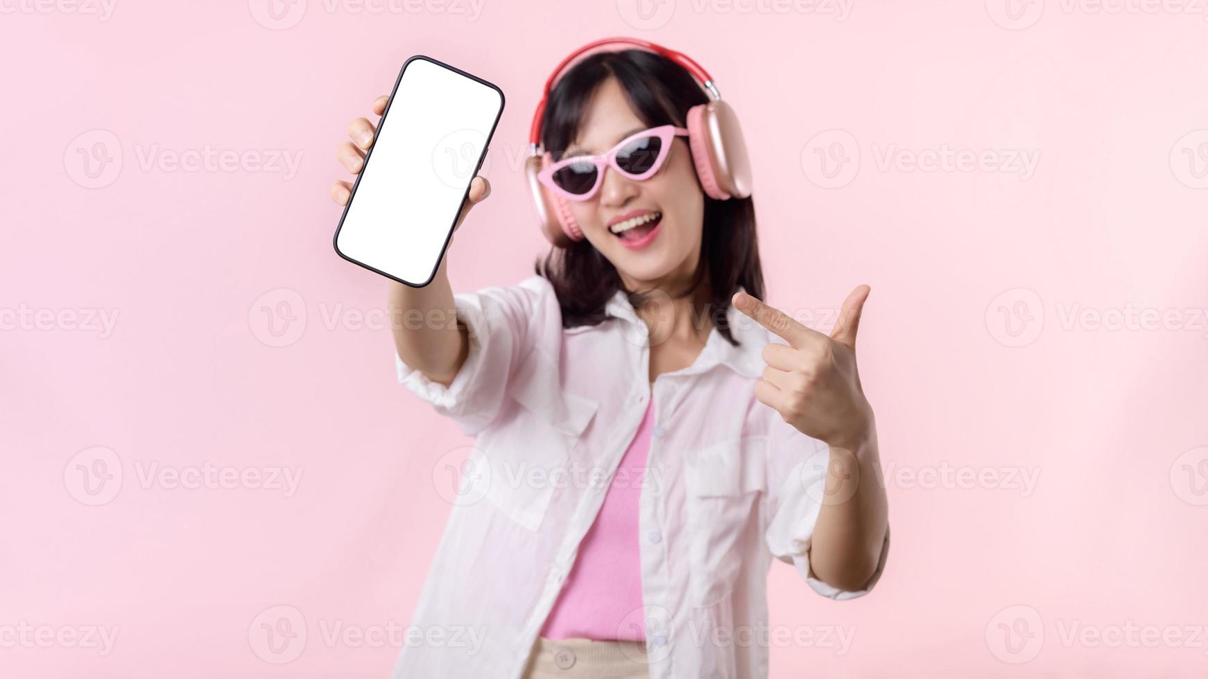 feliz alegre sorridente ásia mulher com sem fio fones de ouvido mostrando em branco tela Móvel telefone ou Novo Smartphone música inscrição propaganda brincar isolado em Rosa estúdio fundo. foto