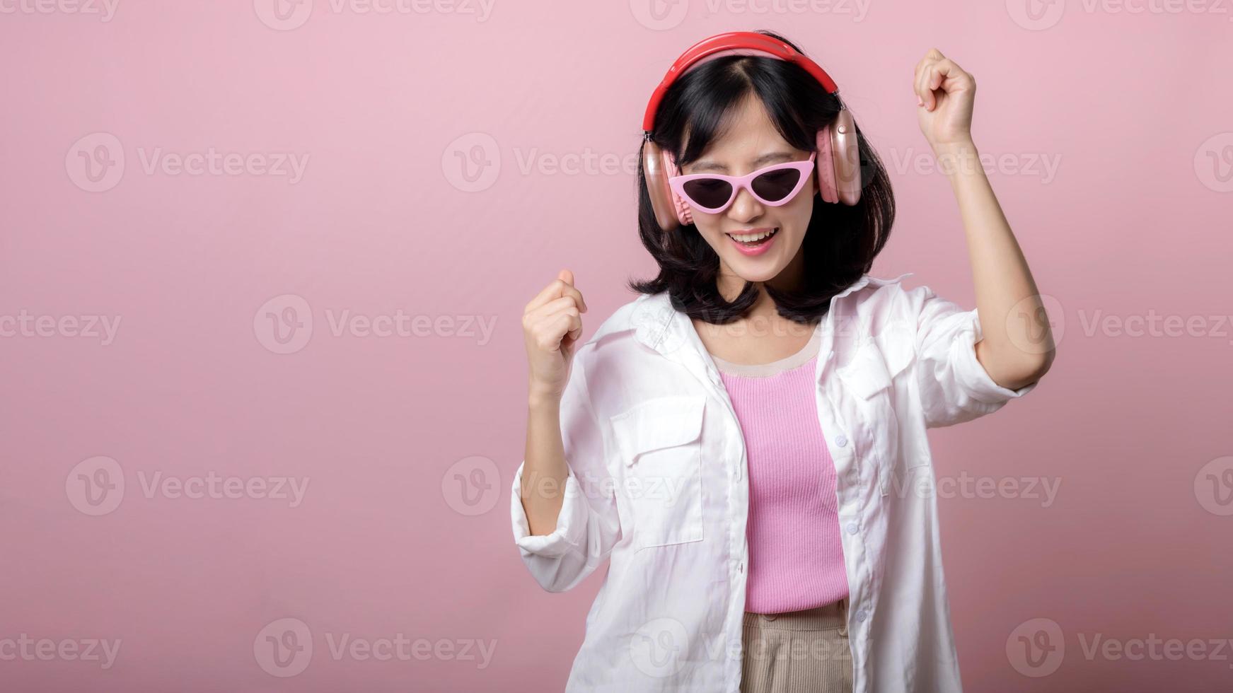 feliz jovem ásia mulher modelo com à moda na moda Sol óculos apreciar ouvindo música de fone de ouvido audio e dançando isolado em Rosa estúdio fundo. tecnologia, menina moda, acessório conceito. foto