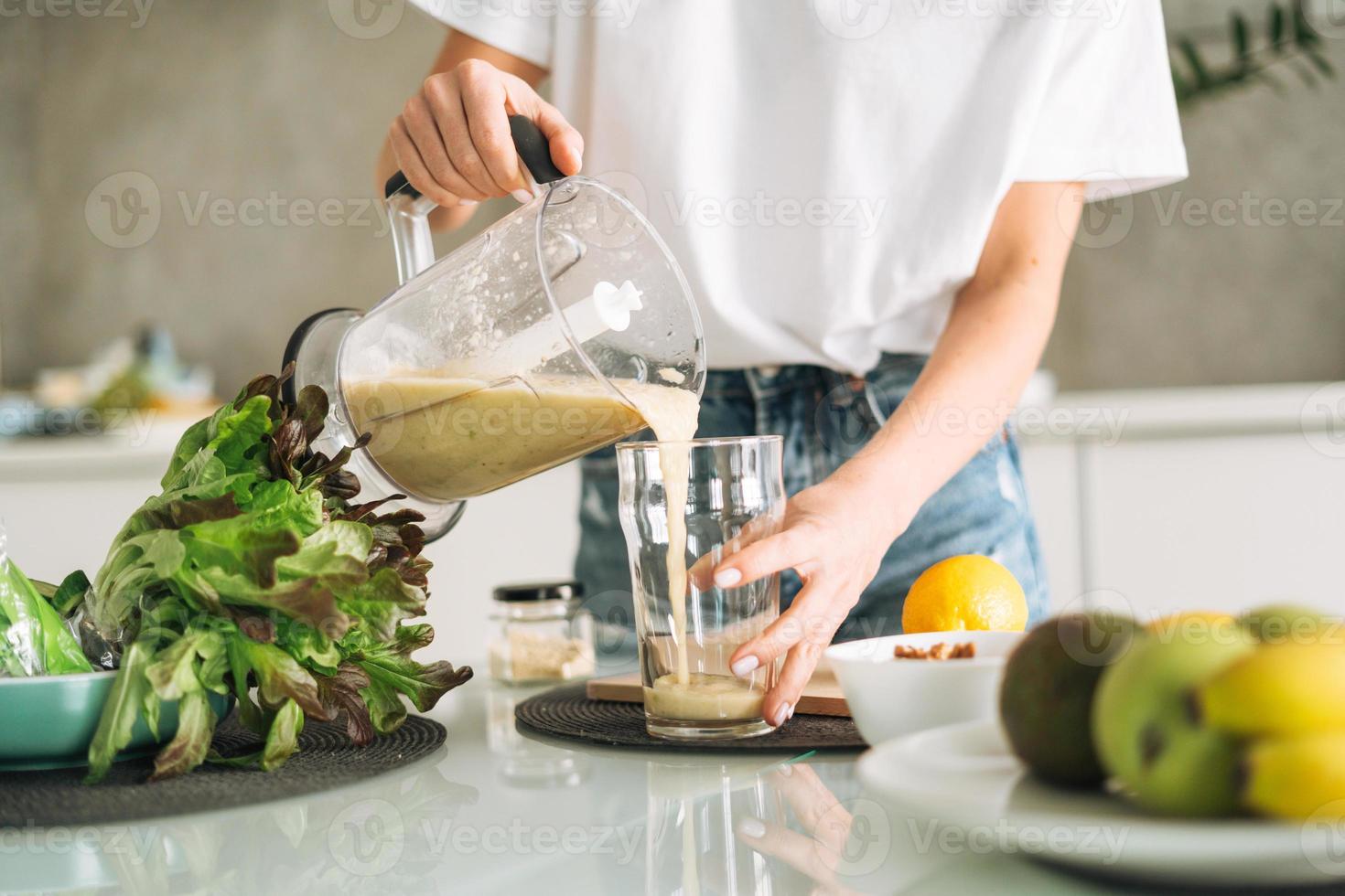 jovem fino mulher dentro branco camiseta e azul jeans cozinhando verde batido saudável Comida dentro cozinha às casa foto