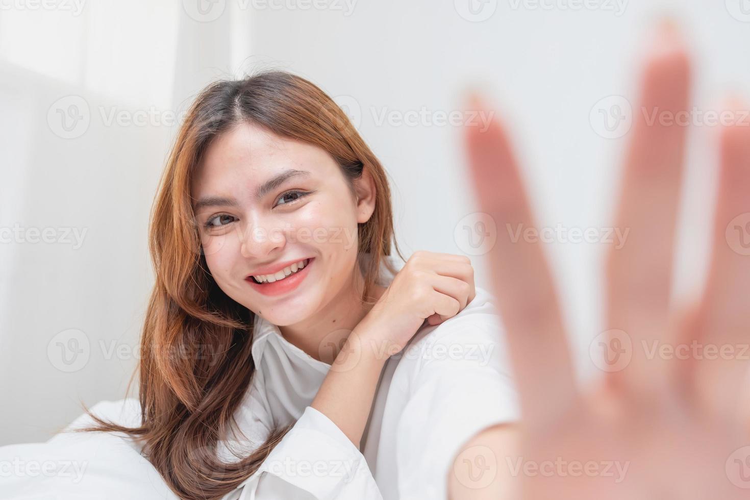 mulher dentro branco camisola acordando acima em final de semana manhã em repouso e relaxante jogando com computador portátil Móvel telefone comendo pão e bebendo chá dentro vidro dentro branco quarto. manhã período de férias conceito. foto