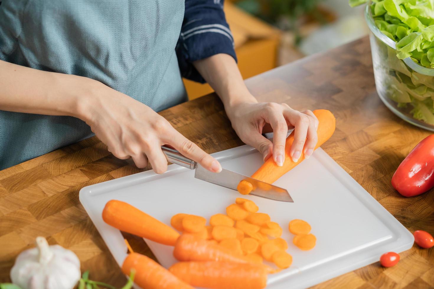 ásia jovem mulher, menina ou dona de casa usando faca, corte cenouras em quadro, em de madeira mesa dentro cozinha lar, preparando ingrediente, receita fresco legumes para cozinhando refeição. saudável Comida pessoas. foto