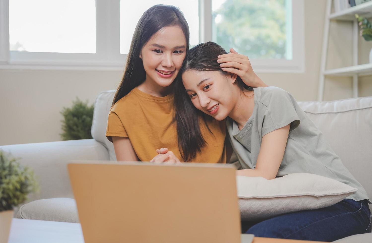 feliz lésbica, prazer ásia jovem dois mulheres, menina gay ou fechar amigo, casal amor abraçar, gastos Boa Tempo junto, usando computador portátil computador para entretenimento em sofá às lar. atividade do lazer. foto