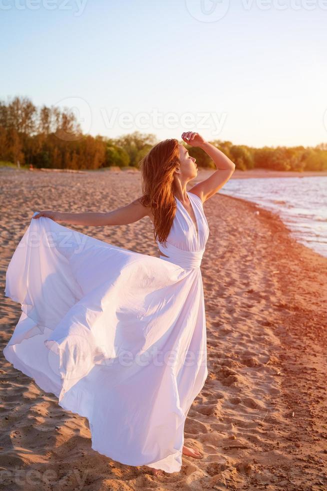 lindo mulher posando em a de praia às pôr do sol dentro uma branco vestir foto