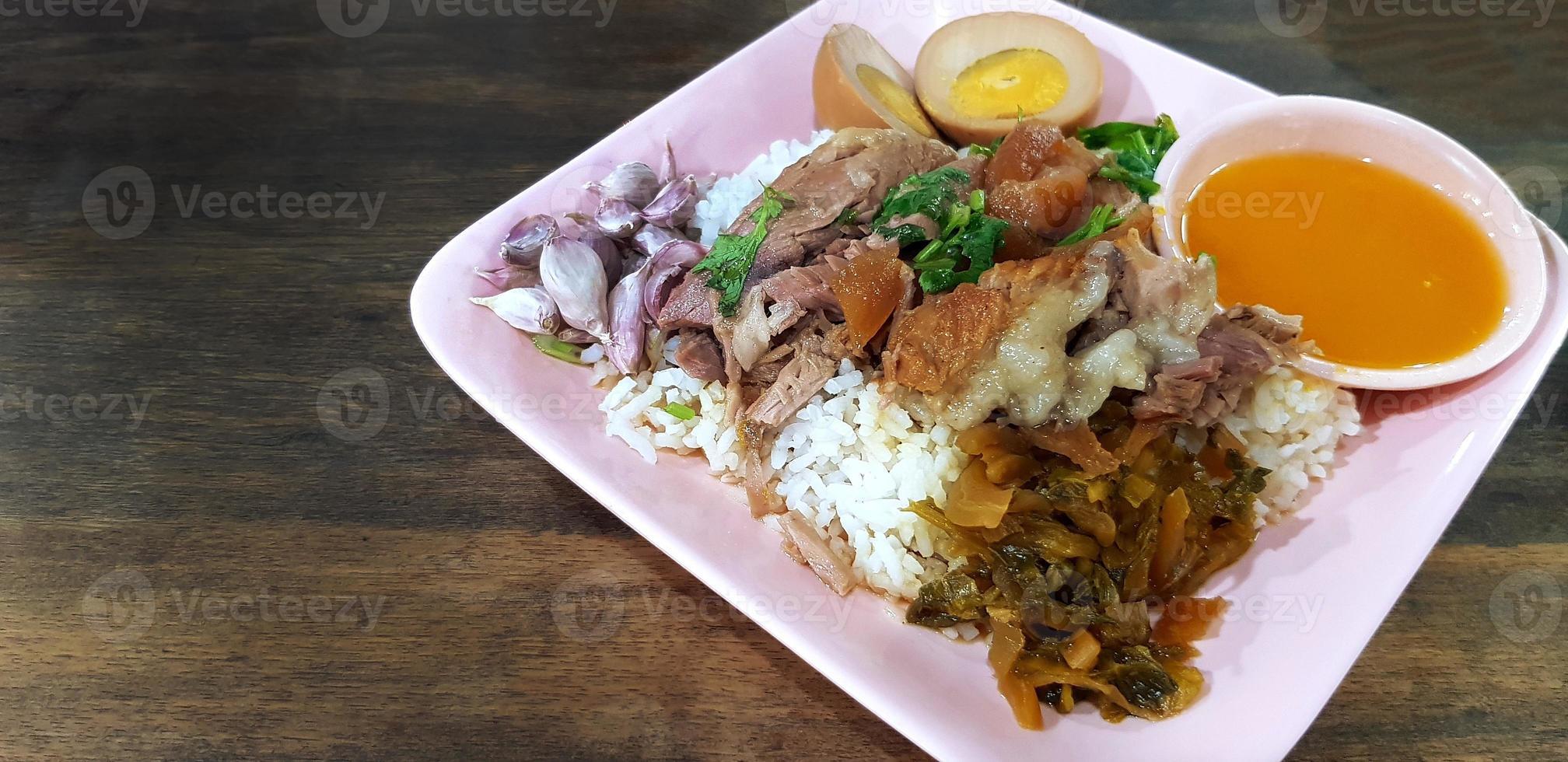 fechar acima cozido carne de porco perna com arroz, alho, fatias do fervido ovo, salmoura, vegetal e azedo molho dentro Rosa prato em Sombrio Castanho de madeira fundo ou mesa com cópia de espaço. tailandês local e rua Comida foto