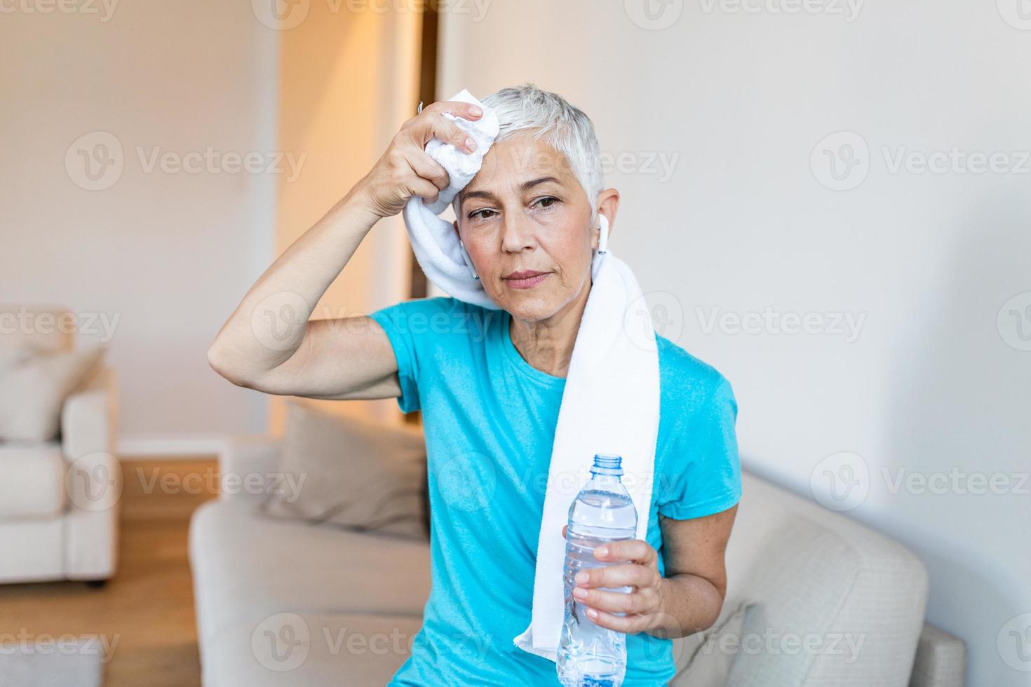 Senior mulher segurando plástico garrafa do água, limpando suor com uma toalha, Exausta depois de a diariamente treinamento. idosos mulher levando uma pausa enquanto exercício às lar. sentado em a sofá e em repouso foto