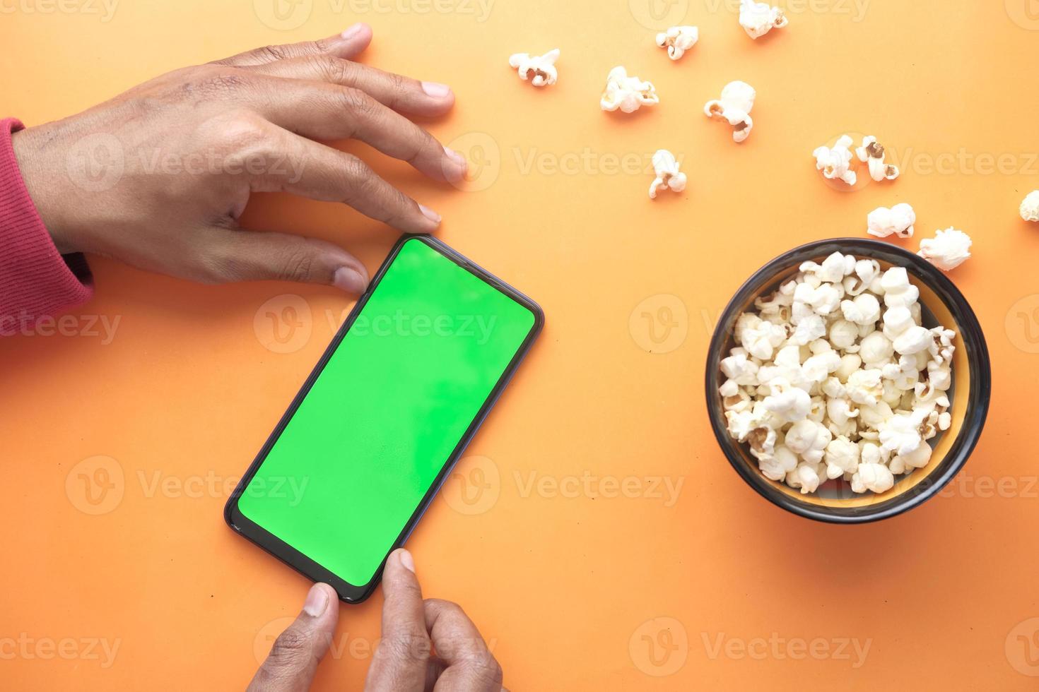 homem usando telefone inteligente e comendo pipoca em fundo laranja foto