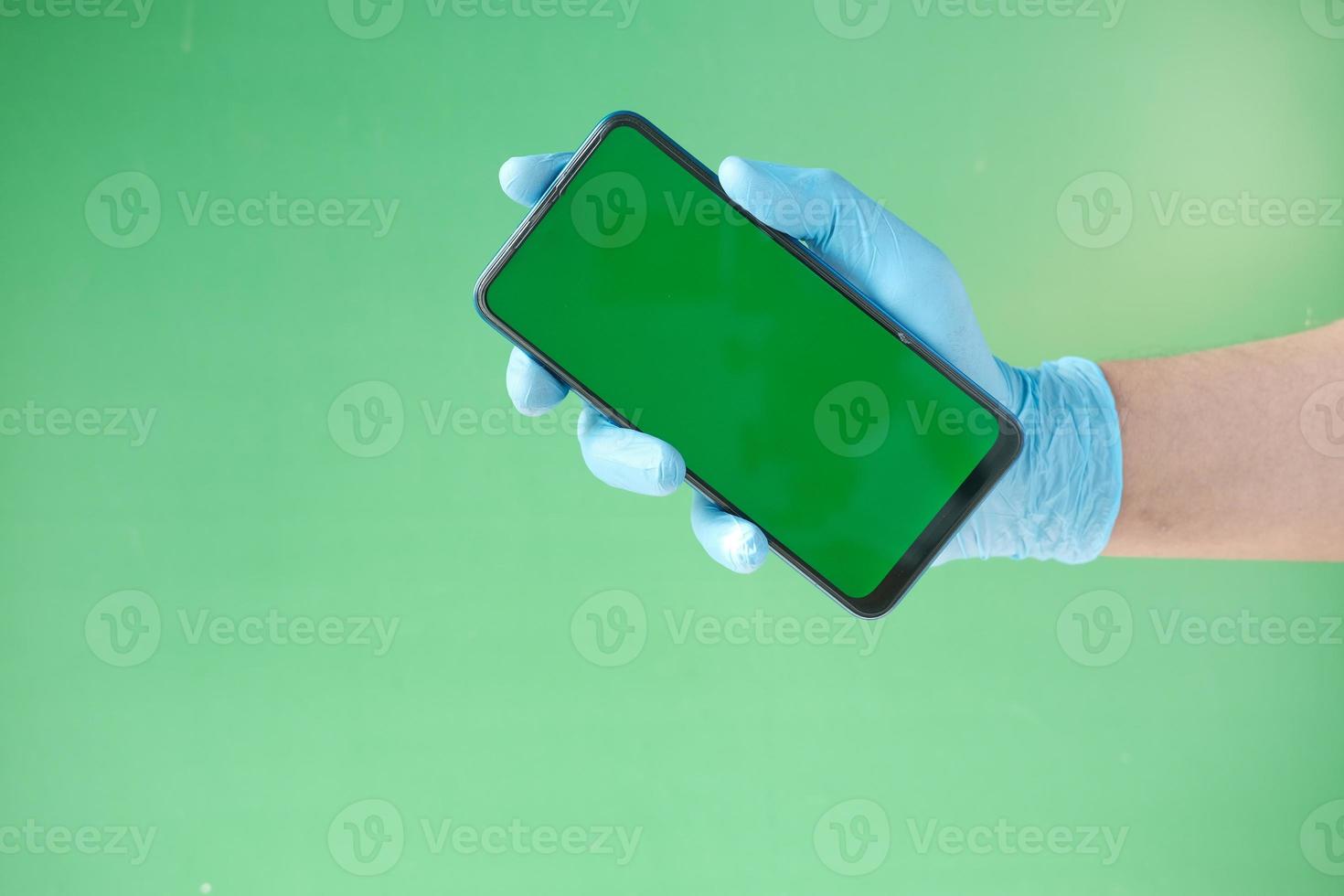 mão em luvas de látex segurando um telefone inteligente contra um fundo verde foto