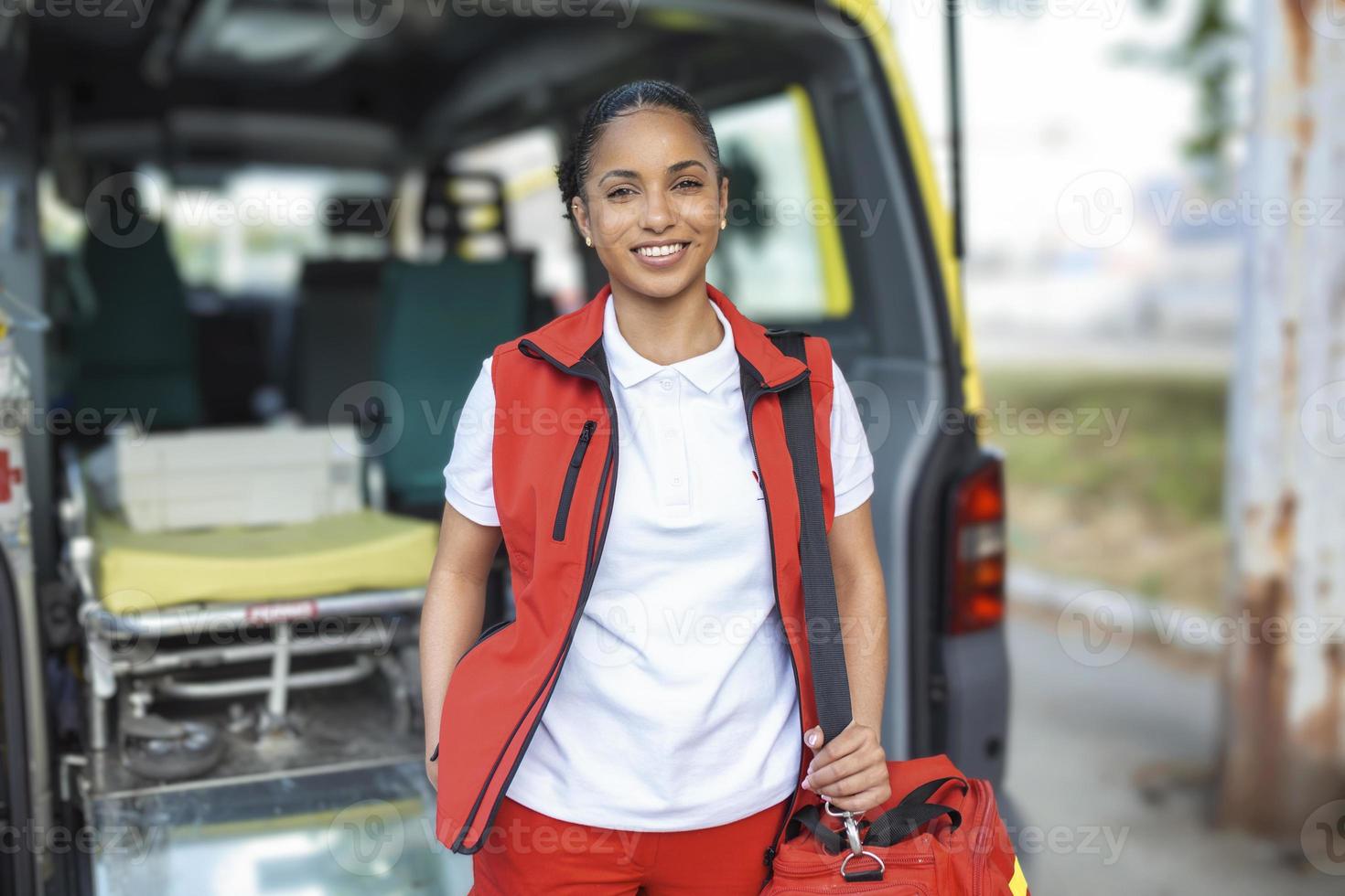 jovem mulher , uma paramédico, em pé às a traseiro do a ambulância, de a aberto portas ela é olhando às a Câmera com uma confiante expressão, sorridente, carregando uma médico trauma saco em dela ombro. foto