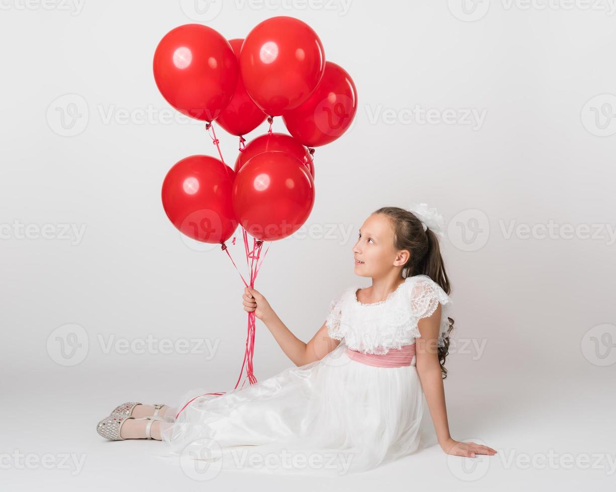 lindo menina vestido dentro grandes branco vestir segurando muitos do vermelho balões dentro mão, olhando acima às bolas foto