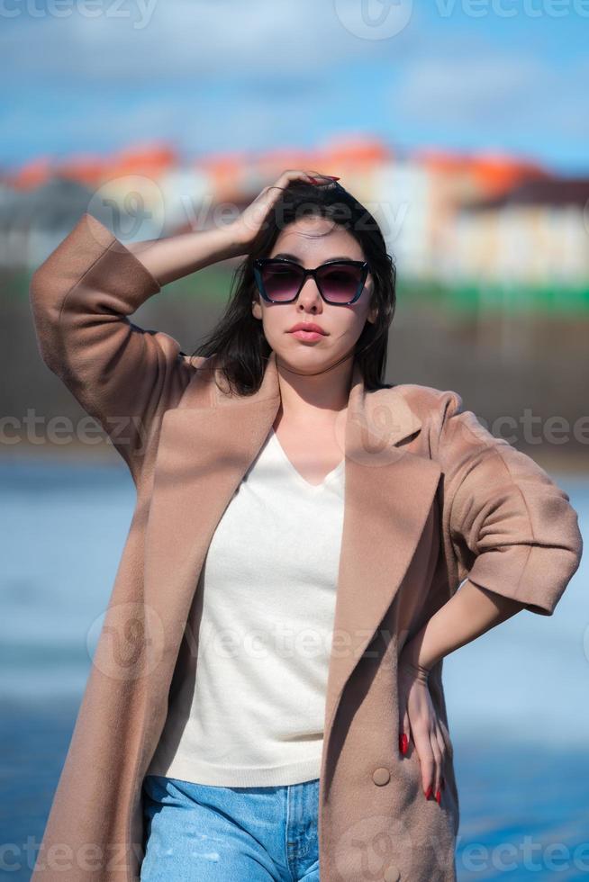 retrato do à moda jovem mulher com grandes cabelo, roliço lábios e oculos de sol, vestido dentro bege casaco foto