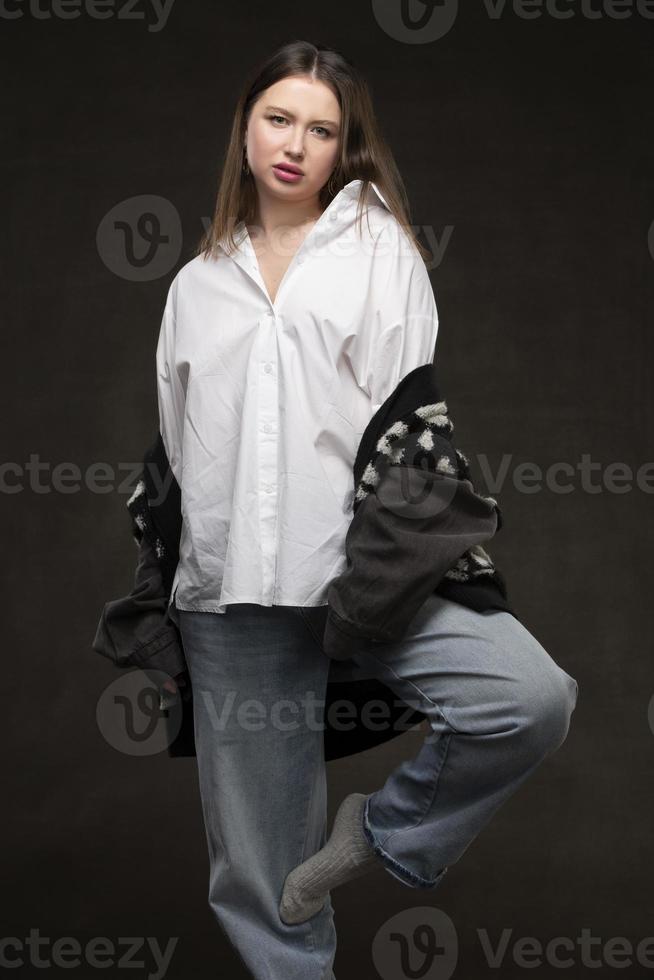 uma lindo menina dentro uma branco camisa e jeans carrinhos em uma cinzento fundo. foto