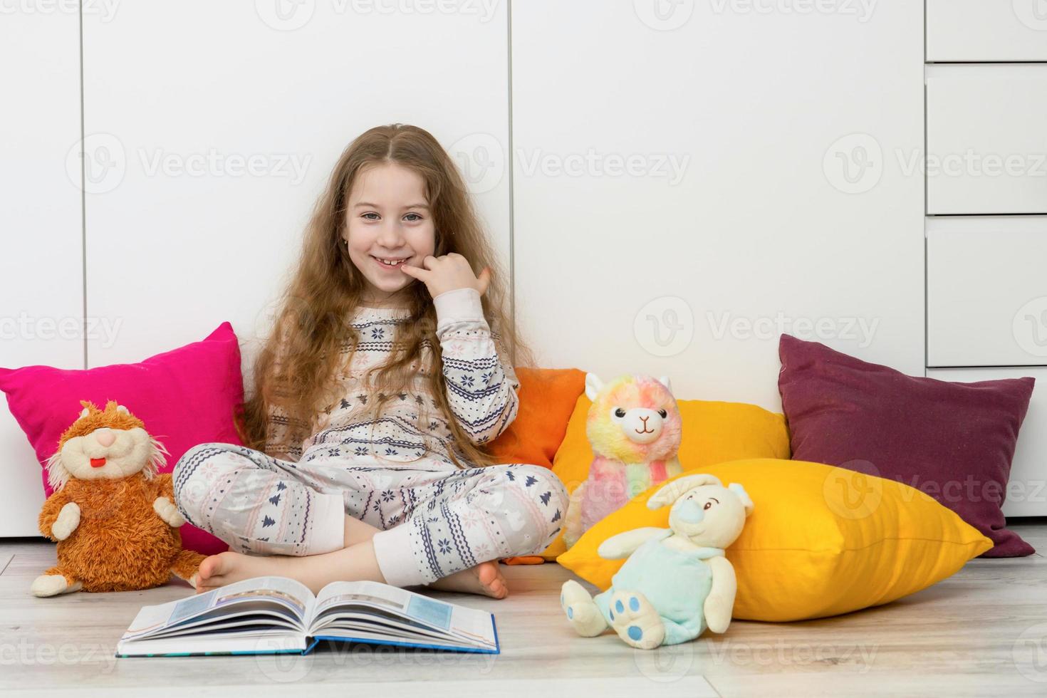 menina dentro pijamas senta em a chão entre colorida almofadas e a aberto livro e risos alegremente foto