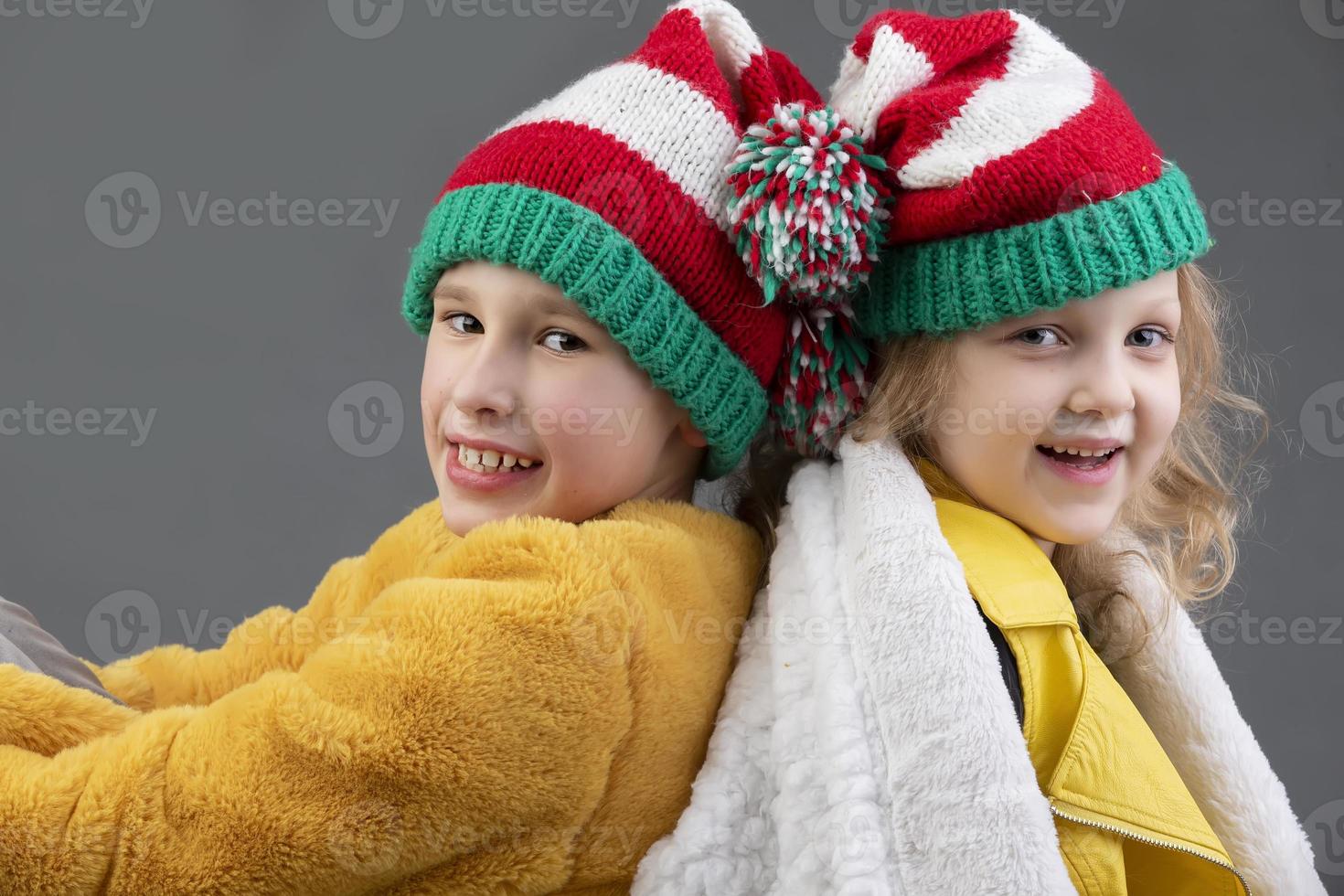 lindo pequeno menina e Garoto dentro tricotado Natal chapéus estão sentado com seus costas para cada de outros e sorridente em uma cinzento fundo. feliz Natal crianças. foto