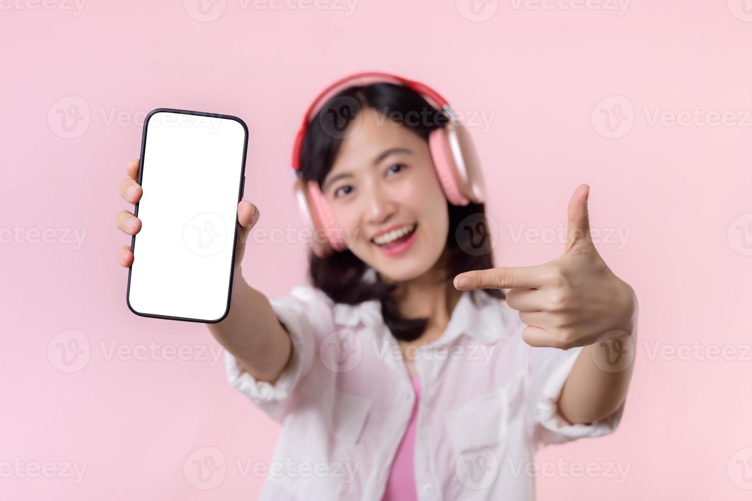 feliz alegre sorridente ásia mulher com sem fio fones de ouvido mostrando em branco tela Móvel telefone ou Novo Smartphone música inscrição propaganda brincar isolado em Rosa estúdio fundo. foto