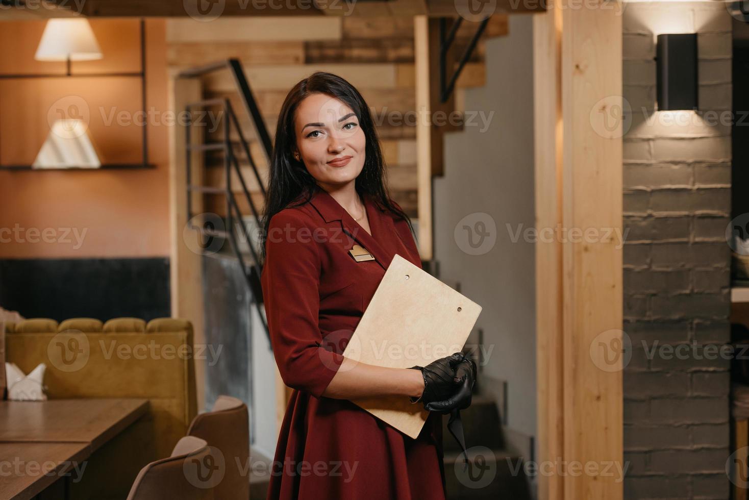 uma sorridente gerente de restaurante com luvas médicas descartáveis segurando um menu de madeira em um restaurante foto