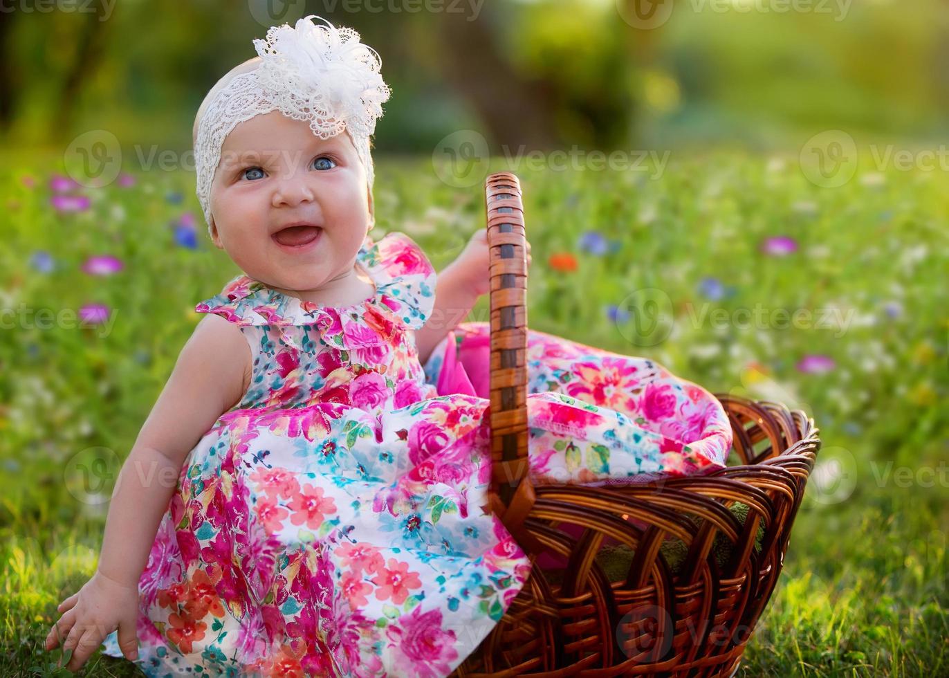 uma lindo pequeno menina senta dentro uma vime cesta e risos alegremente em uma flor Prado. lindo crianças. verão andar do uma fofa bebê. feliz criança acima para uma ano. foto