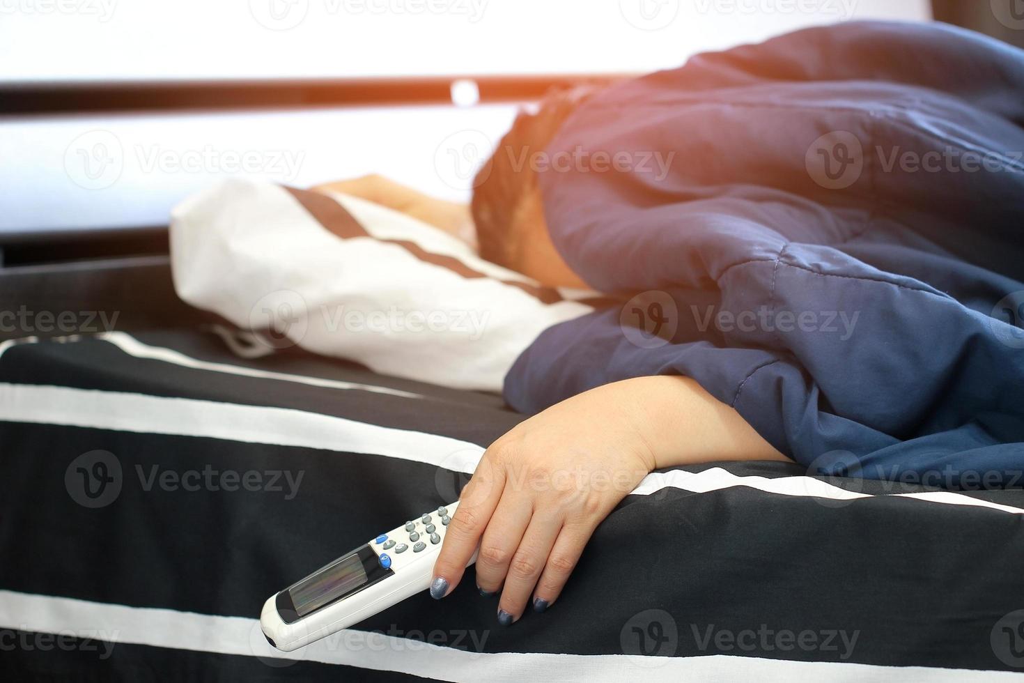 mulher dormindo na cama em um dia quente e ensolarado segurando um controle remoto para o ar condicionado para ajustar a temperatura de acordo com o corpo e para uma boa saúde foto