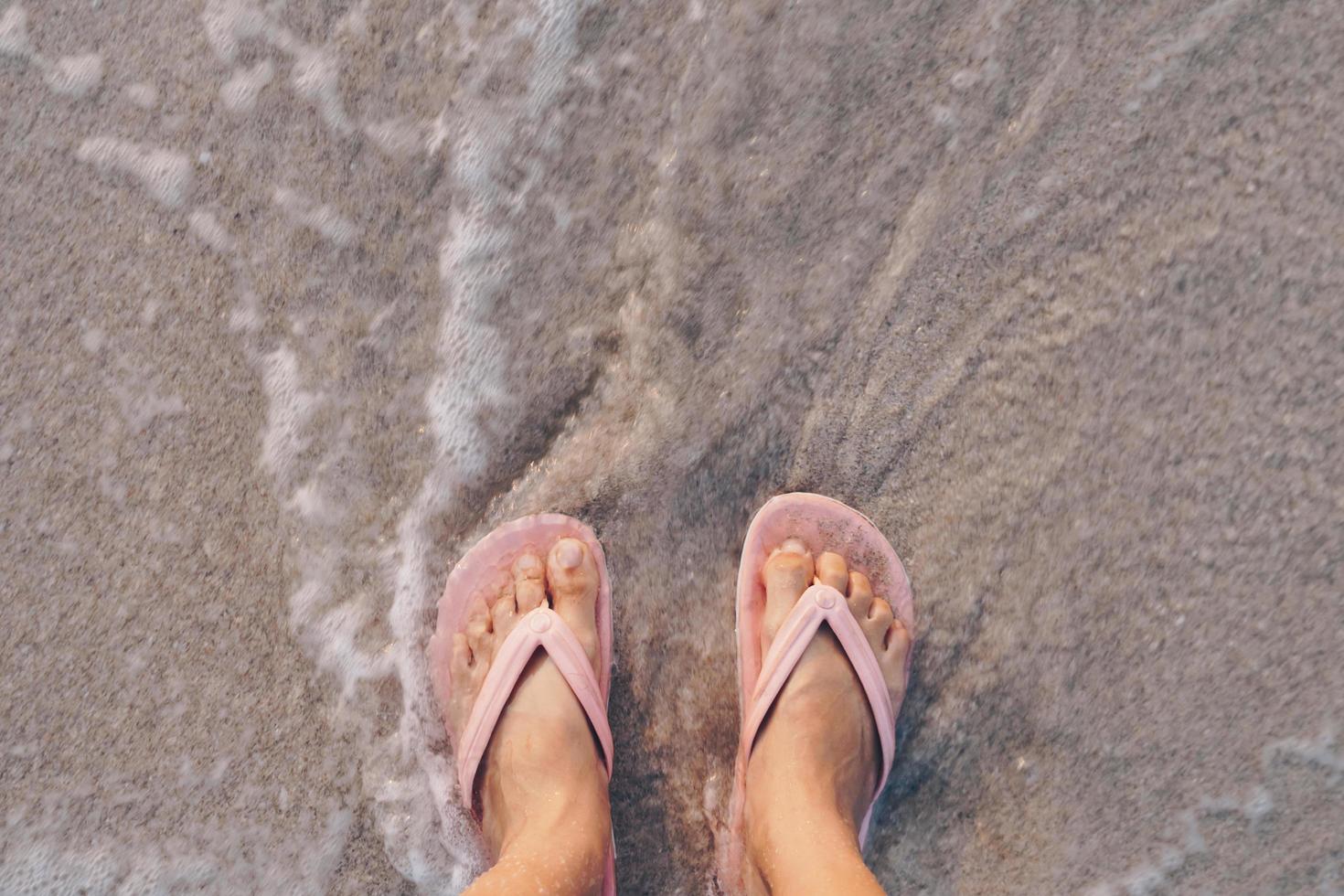 vista superior dos pés de uma mulher de chinelos em uma praia arenosa com as ondas do mar foto