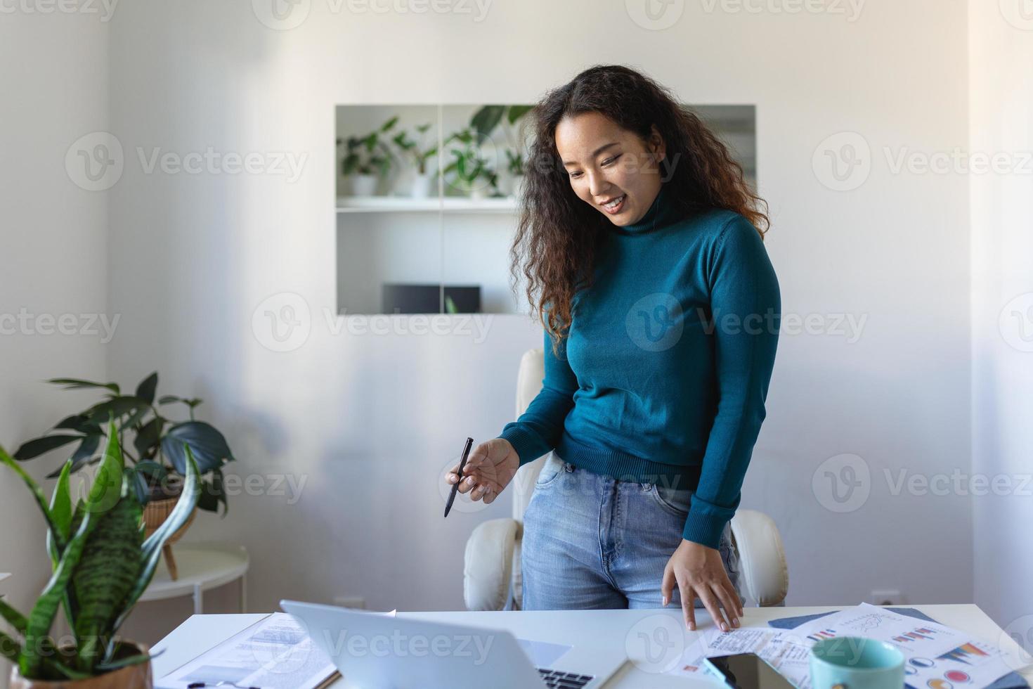 lindo sorridente ásia empresária lendo alguma coisa em uma computador portátil enquanto inclinado em a mesa dentro a escritório foto