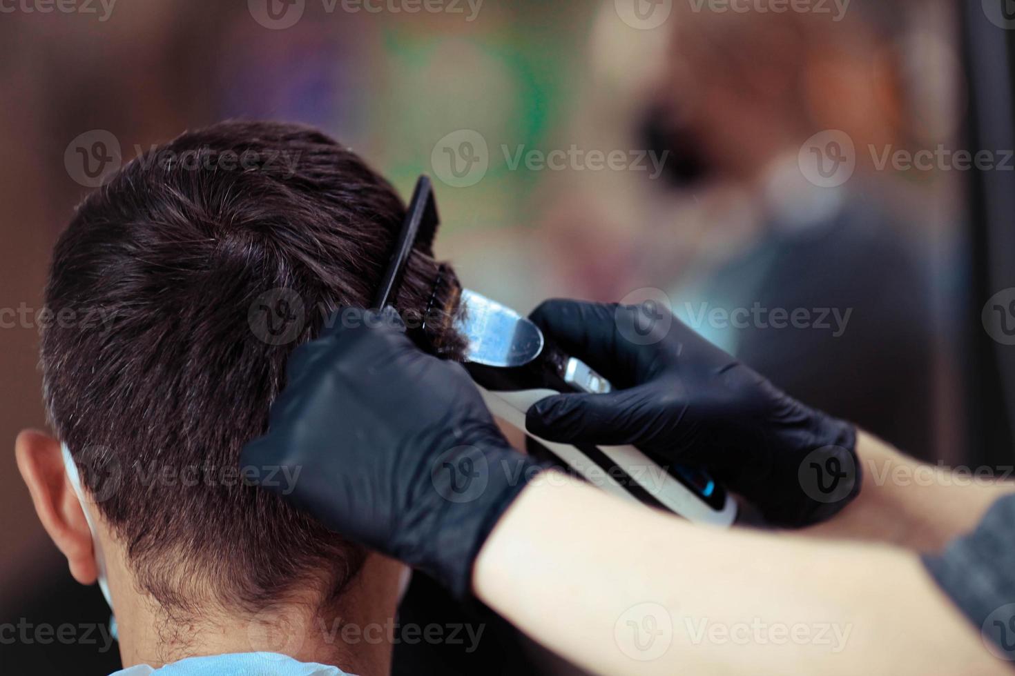 uma cabeleireiro com segurança medidas para COVID-19, detém uma navalha e uma corte de cabelo para uma homem, cortes dele cabelo com borracha luvas dentro uma beleza salão foto