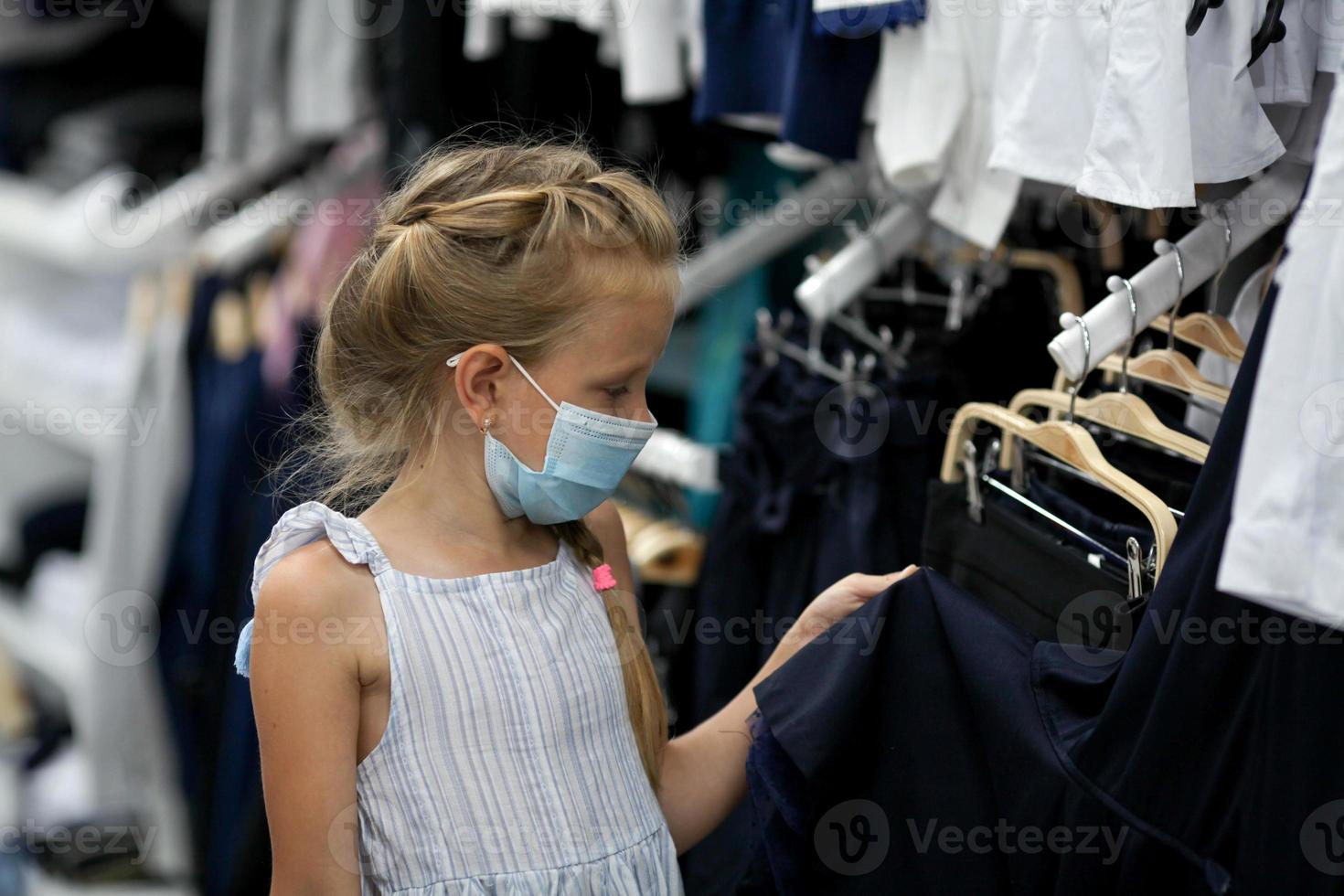 pequeno menina dentro uma médico mascarar escolhe escola uniforme loja. preparando para escola depois de quarentena cobiçado - 19. protetora mascarar para a prevenção do coronavírus. foto