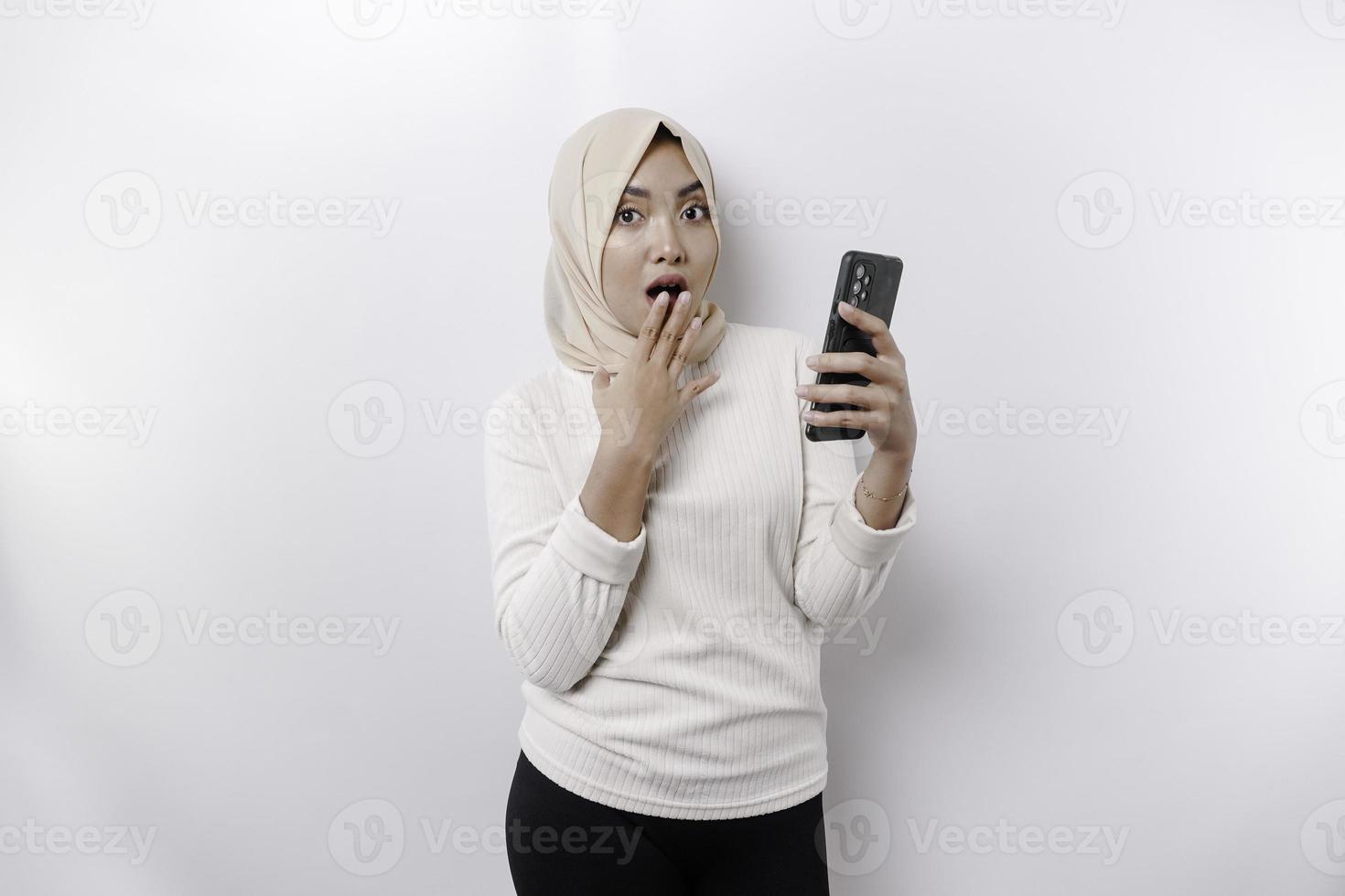 chocado ásia mulher vestindo lenço de cabeça, segurando dela telefone, isolado de branco fundo foto