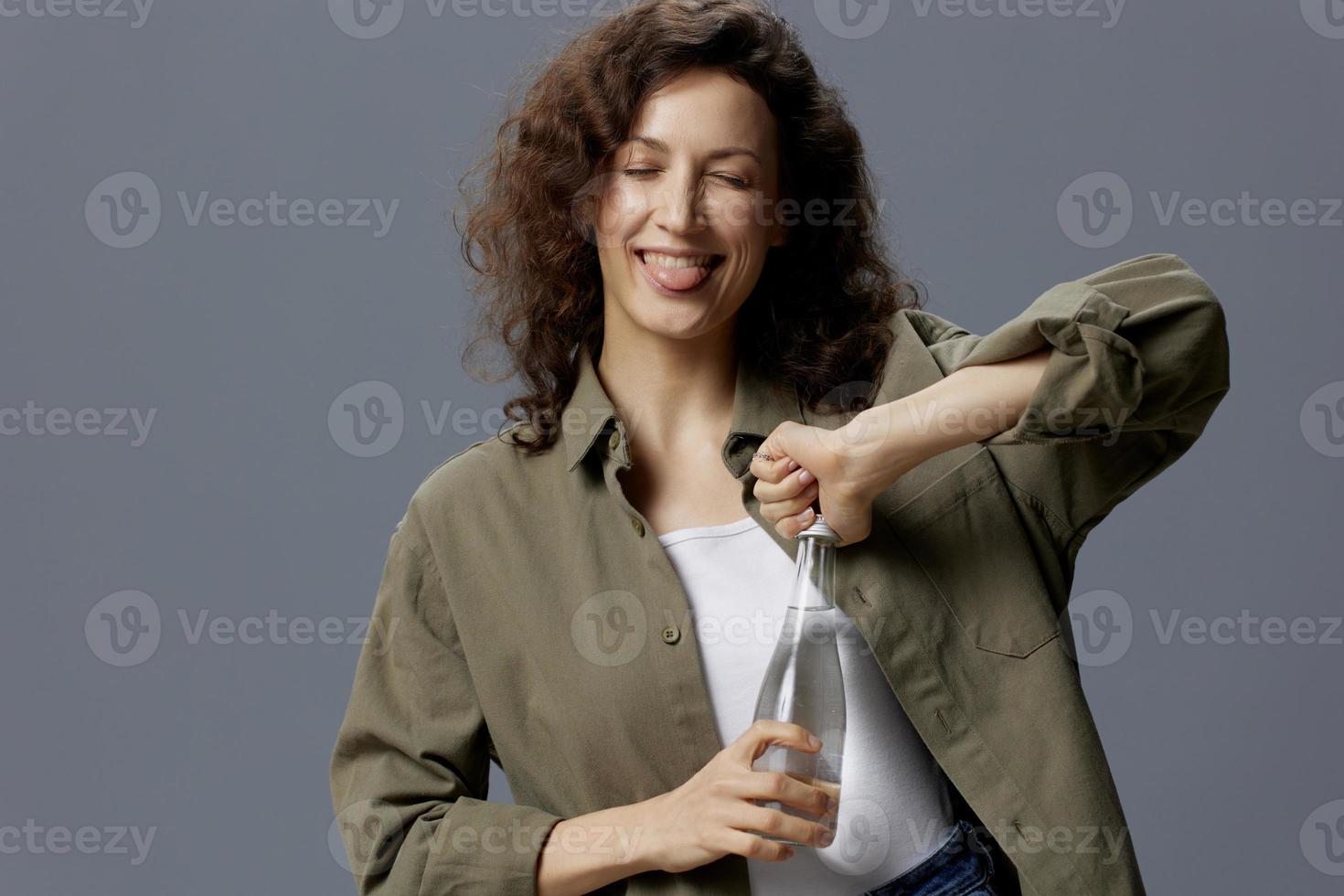 feliz sorridente encaracolado lindo mulher dentro casual cáqui verde camisa aguarde aberto água garrafa Gravetos língua Fora posando isolado em cinzento azul fundo. saudável estilo de vida. água é vida conceito. cópia de espaço foto