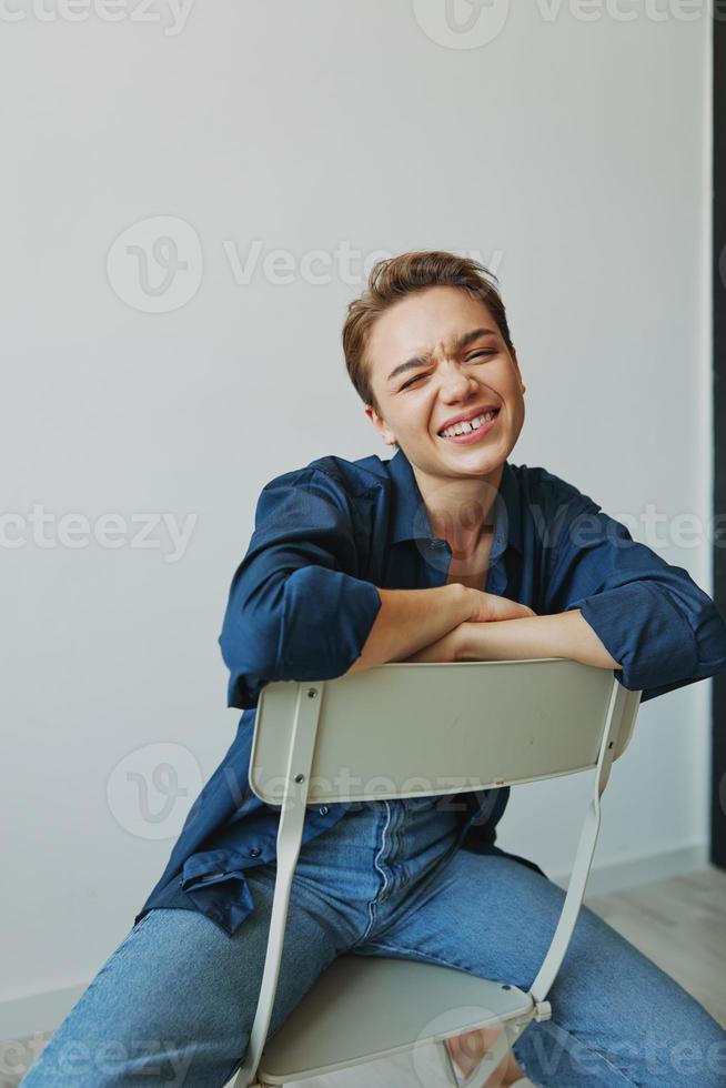 uma jovem mulher sentado dentro uma cadeira às casa sorridente com dentes com uma curto corte de cabelo dentro jeans e uma jeans camisa em uma branco fundo. menina natural poses com não filtros foto