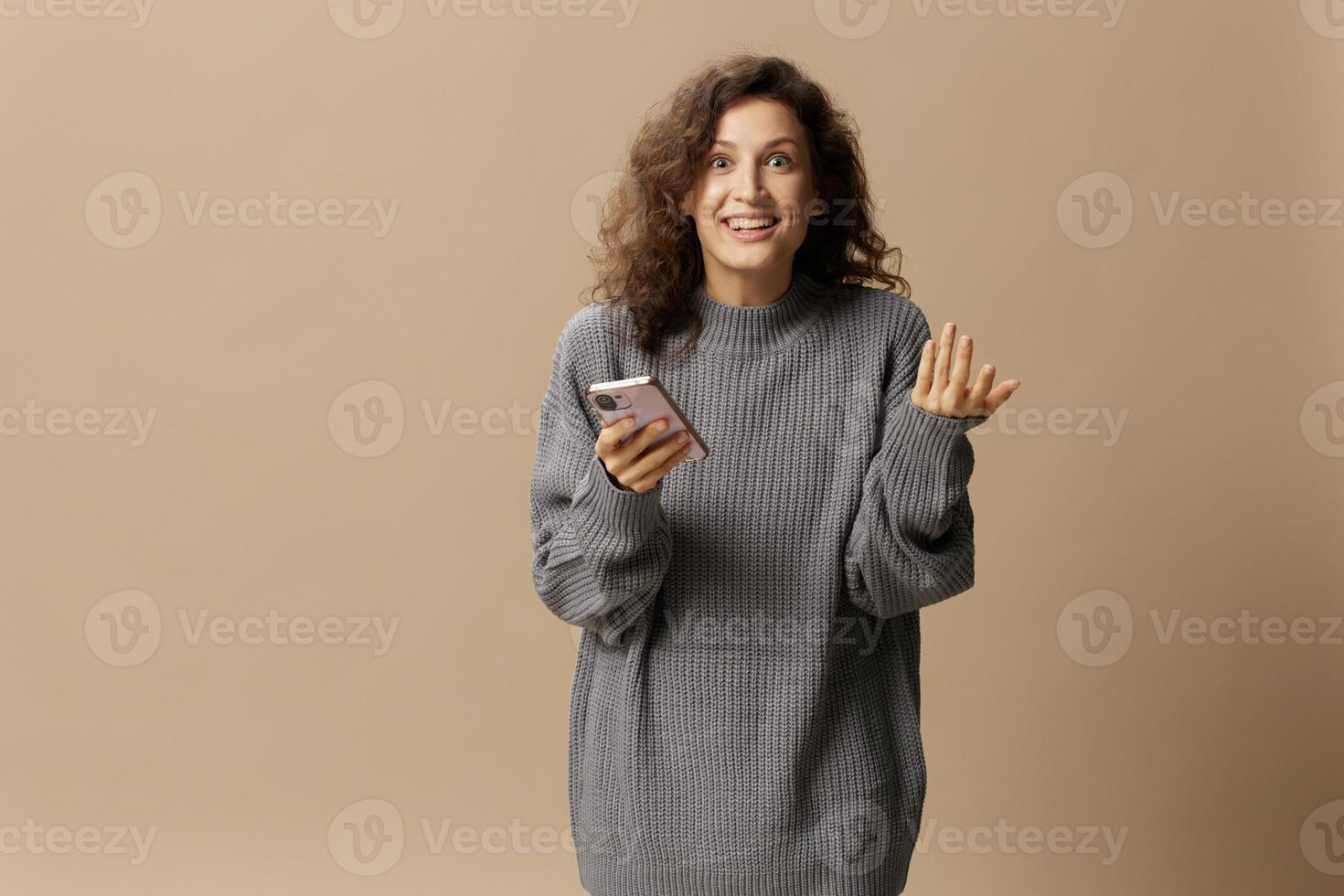 animado feliz encaracolado lindo senhora dentro cinzento casual suéter pegue legal notícia a partir de mensagem posando isolado em sobre bege pastel fundo. social meios de comunicação, rede, distância comunicação conceito. cópia de espaço foto