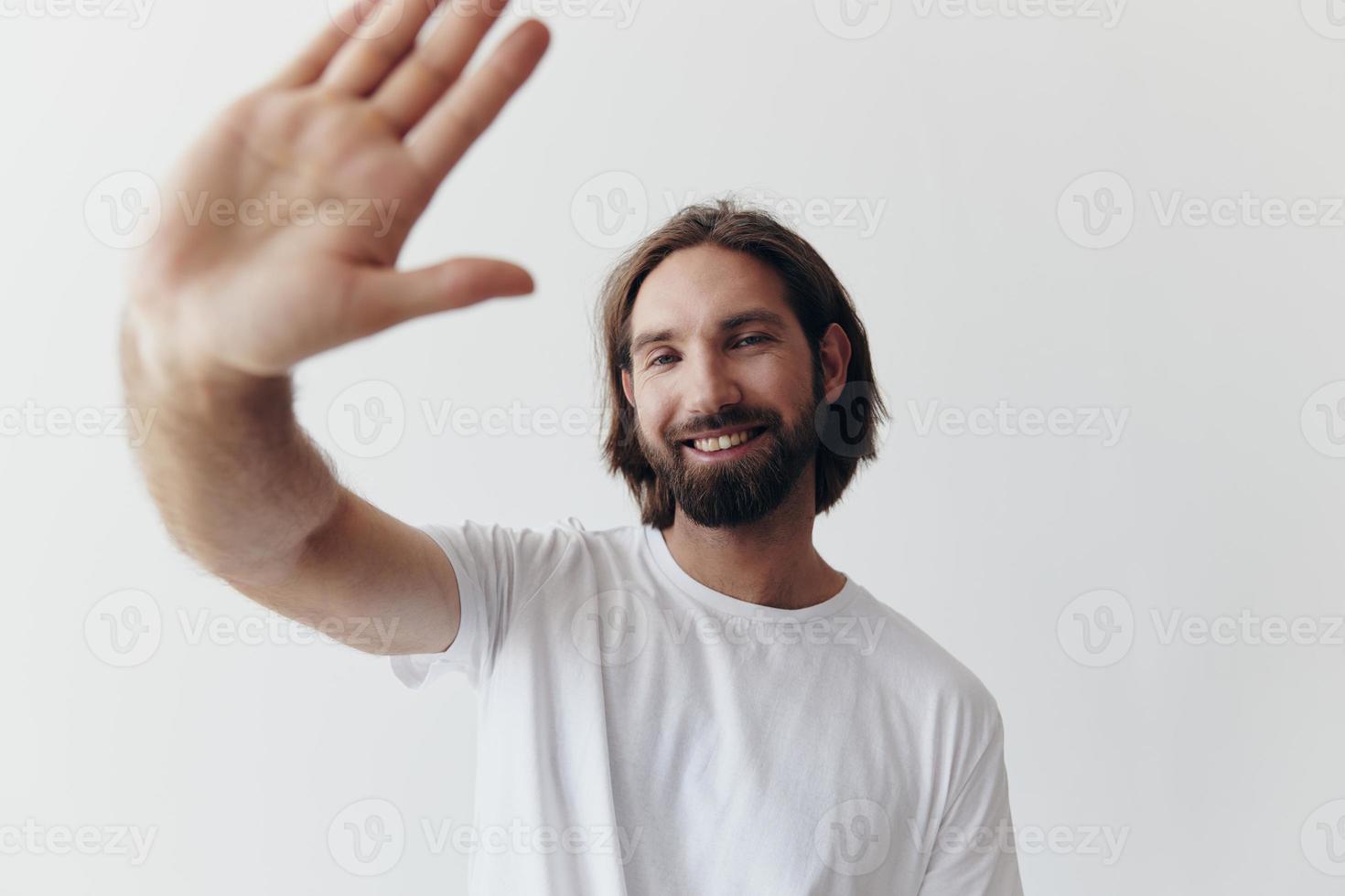 feliz adulto homem com uma barba sorrisos e puxa a cozinhar para dentro a Câmera ouvindo para música dentro fones de ouvido dentro uma angustiado camiseta em uma branco isolado fundo foto