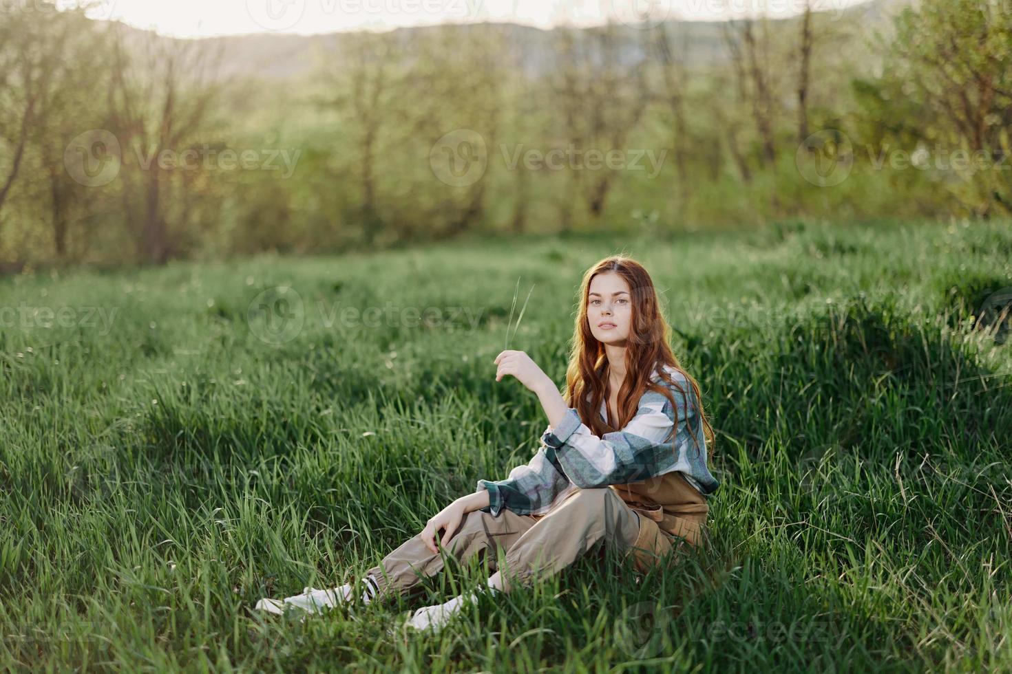 uma jovem lindo mulher senta em a verde Relva dentro a parque e parece Fora para dentro a configuração Sol foto