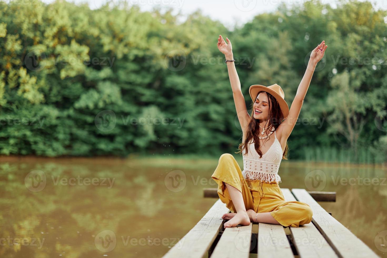 hippie eco-ativista mulher viajante senta em uma ponte de uma lago com dela braços estendido com uma chapéu e sorridente Atenciosamente foto