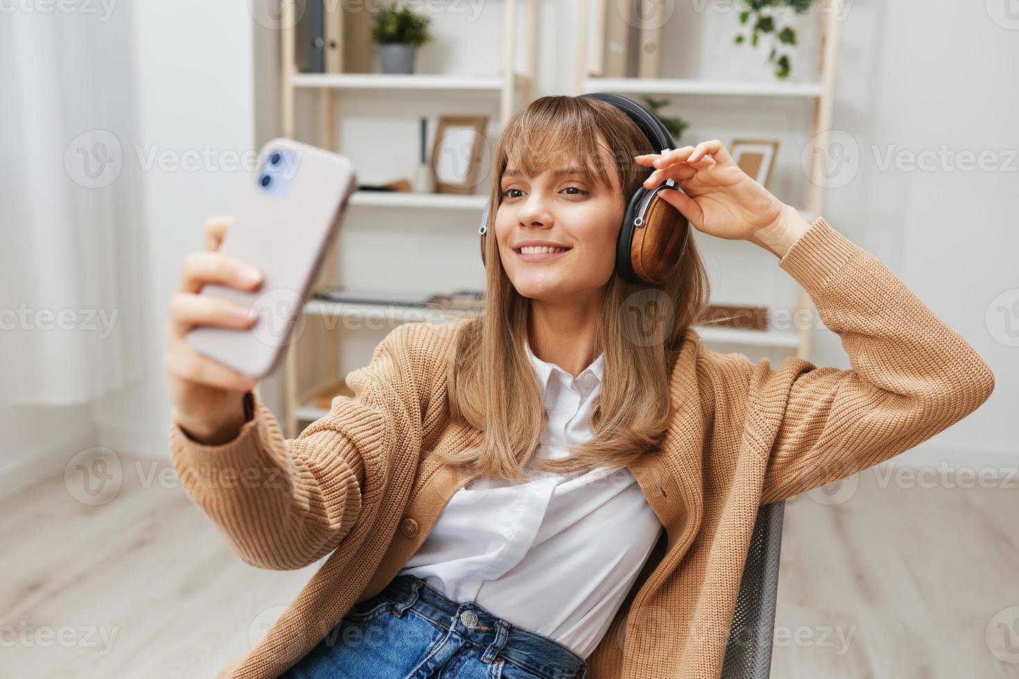 alegre jovem Loiras senhora dentro caloroso suéter dentro fones de ouvido ouço fav canções fazendo selfie vídeo ligar sentado dentro poltrona às moderno casa interior. música Tempo relaxante legal lista de reprodução conceito. cópia de espaço foto