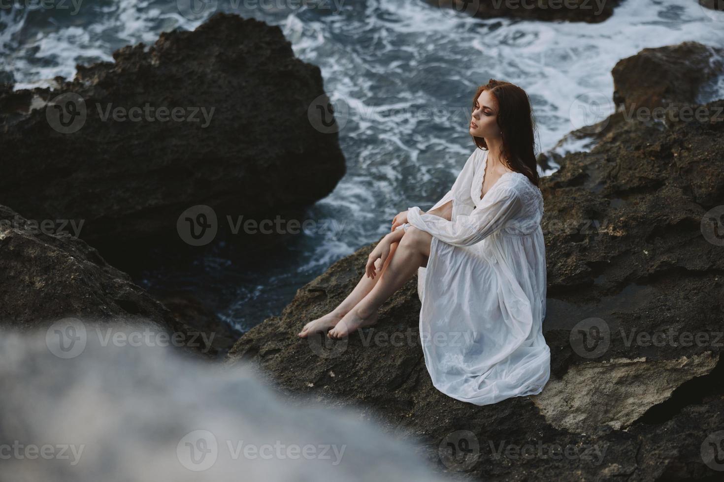 uma mulher dentro uma branco Casamento vestir é sentado descalço em uma onda penhasco inalterado foto