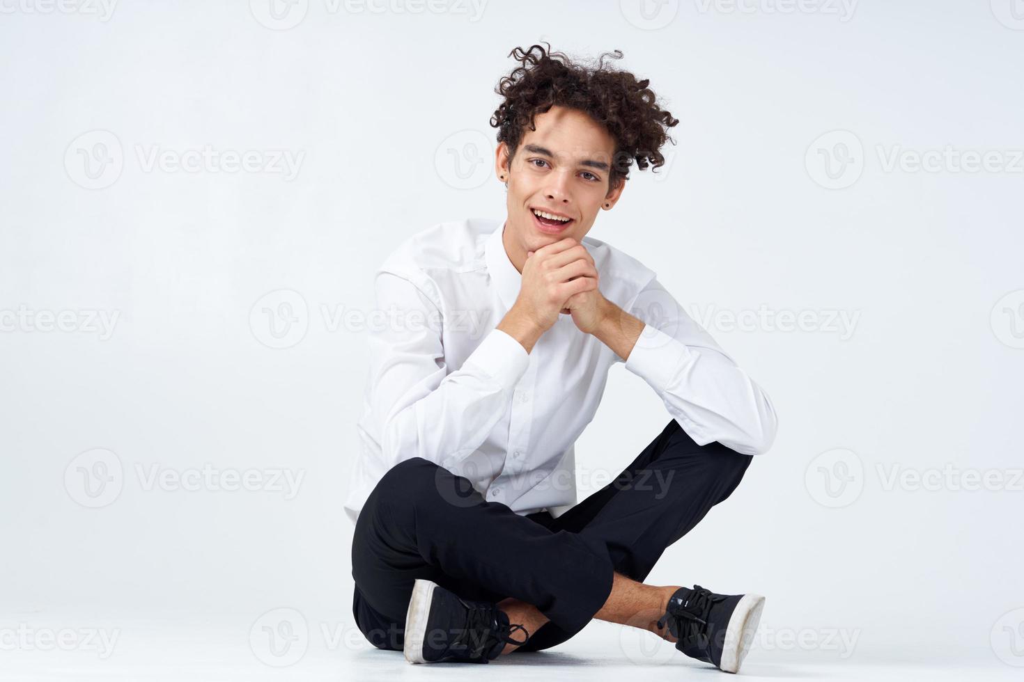 cara com encaracolado cabelo dentro uma branco camisa, tênis e calças senta em a chão foto