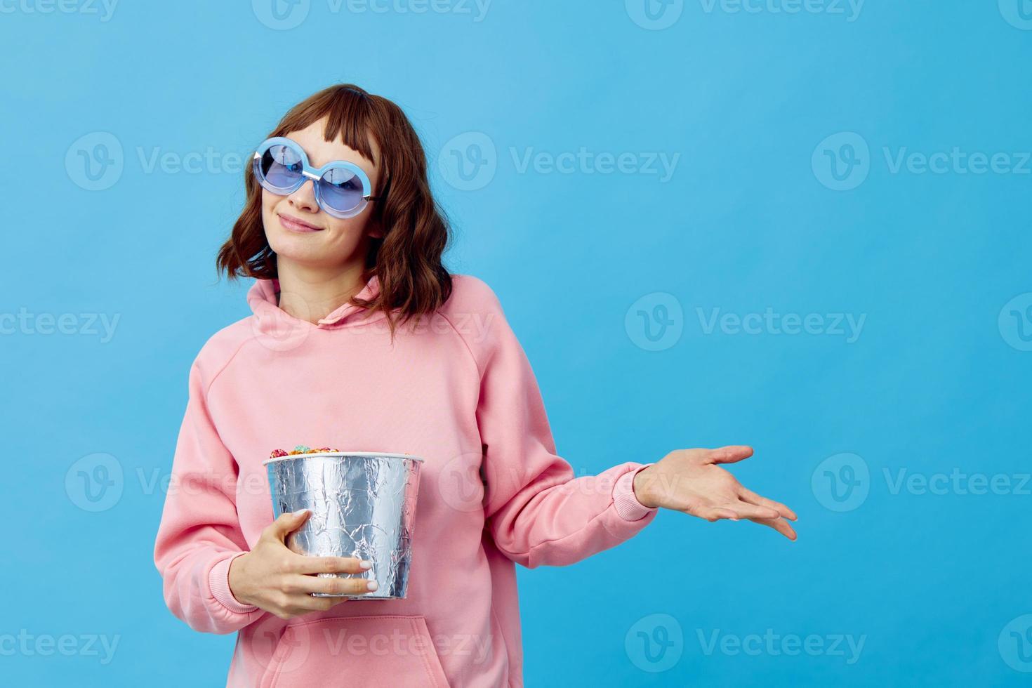 tarde filme conceito. confuso fofa ruiva senhora dentro Rosa moletom com capuz oculos de sol com Pipoca posando isolado em azul estúdio fundo. cópia de espaço bandeira. moda cinema. entretenimento oferta foto