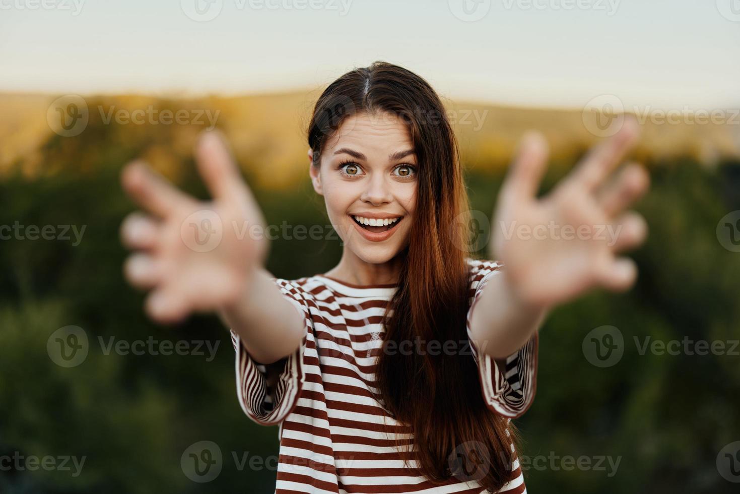 uma mulher sorrisos e puxa dela mãos para a Câmera fechar-se dentro natureza com uma Visão do a montanhas. feliz viagem estilo de vida Segue mim foto