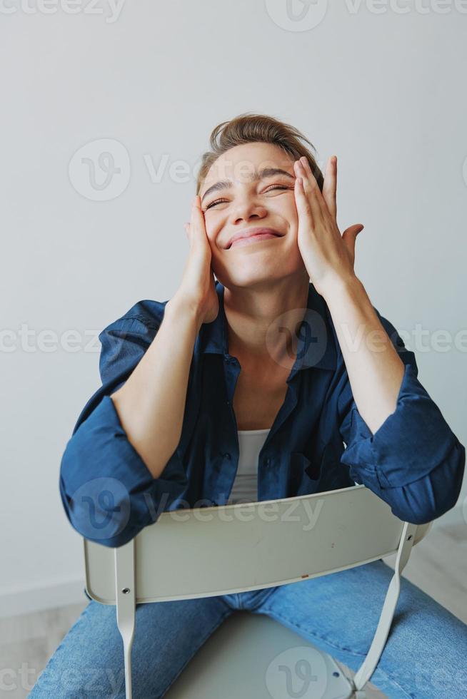 uma jovem mulher sentado dentro uma cadeira às casa sorridente com dentes com uma curto corte de cabelo dentro jeans e uma jeans camisa em uma branco fundo. menina natural poses com não filtros foto