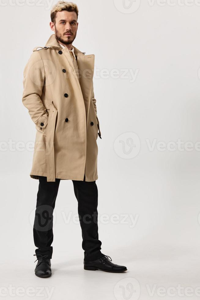 uma delgado cara dentro uma bege casaco detém dele mãos dentro dele bolsos e Sombrio calças modelo foto