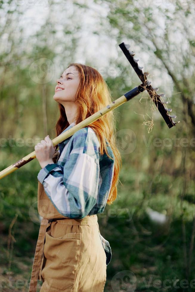 uma mulher agricultor dentro trabalhos roupas e a avental trabalho ao ar livre dentro natureza e detém uma ancinho para reunir Relva foto