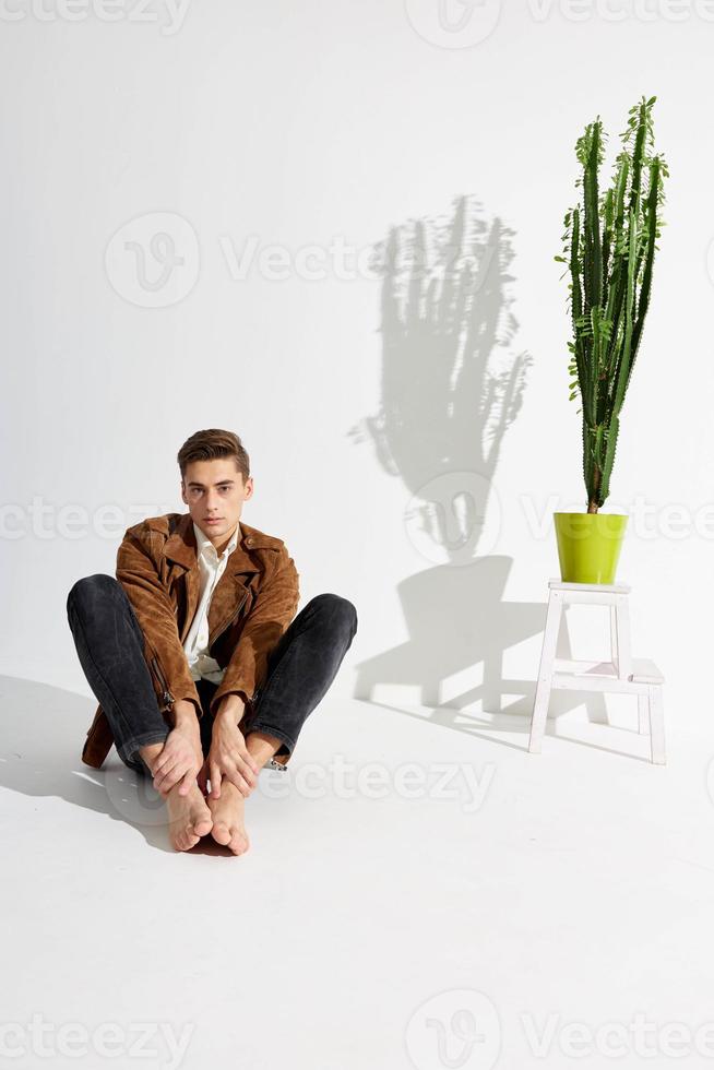 uma homem dentro calças e uma Castanho Jaqueta senta em a chão dentro uma quarto perto uma vaso de flores foto
