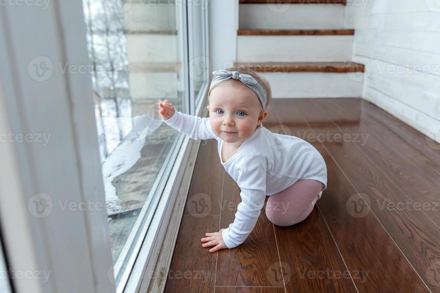 garotinha rastejante de um ano de idade sentada no chão na sala de estar de luz brilhante perto da janela sorrindo e rindo foto