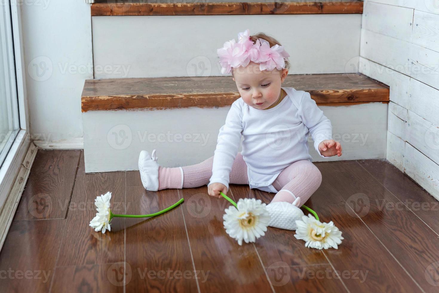 pequeno bebê menina vestindo Primavera guirlanda localização em chão dentro brilhante luz vivo quarto perto janela e jogando com gerbera flores foto