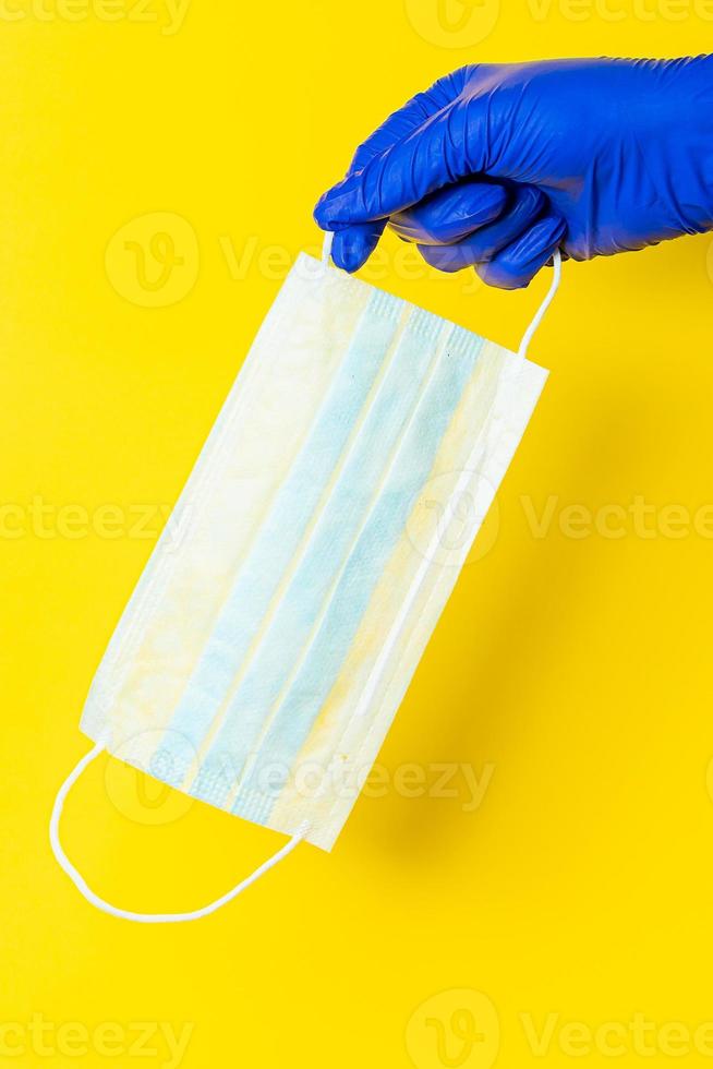 uma mão em uma luva de látex azul segura uma máscara cirúrgica em um fundo amarelo foto