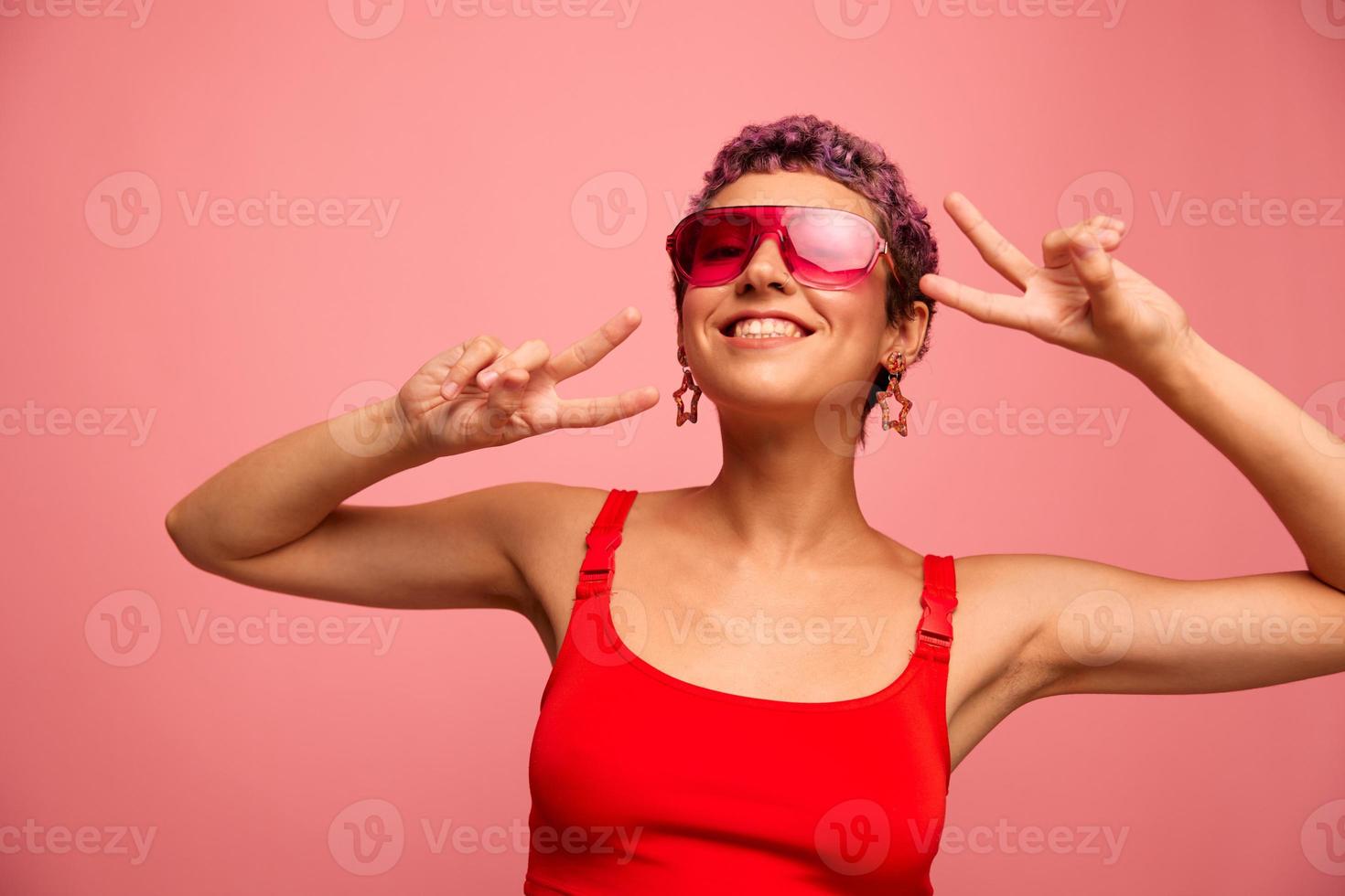 moda retrato do uma mulher com uma curto corte de cabelo dentro colori oculos de sol com incomum acessórios com brincos sorrisos em uma Rosa brilhante fundo mostra dela mãos para a Câmera foto